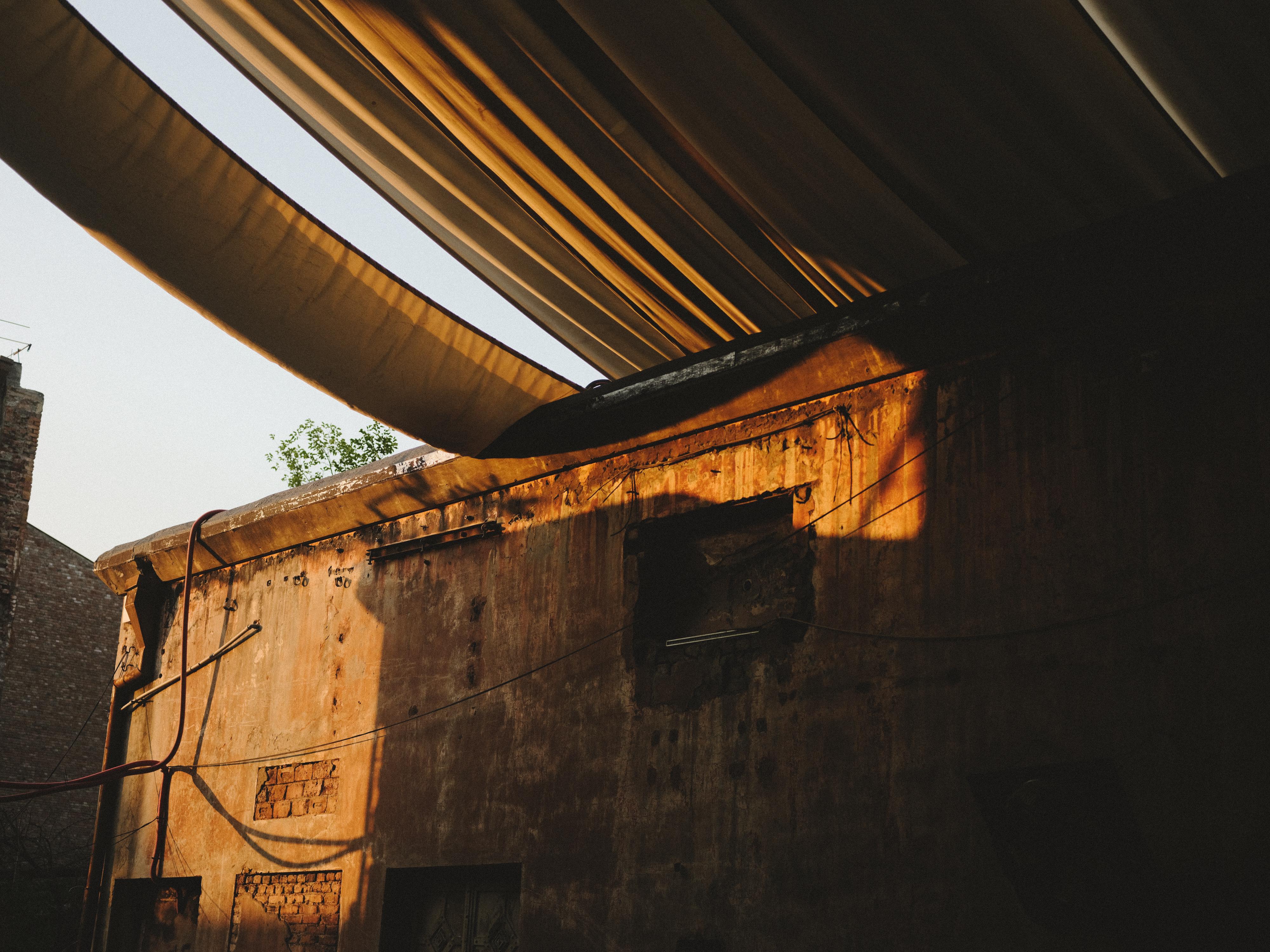 Damaged building interior with partially collapsed corrugated metal roof, exposed wooden beams, and sunlight streaming through gaps.