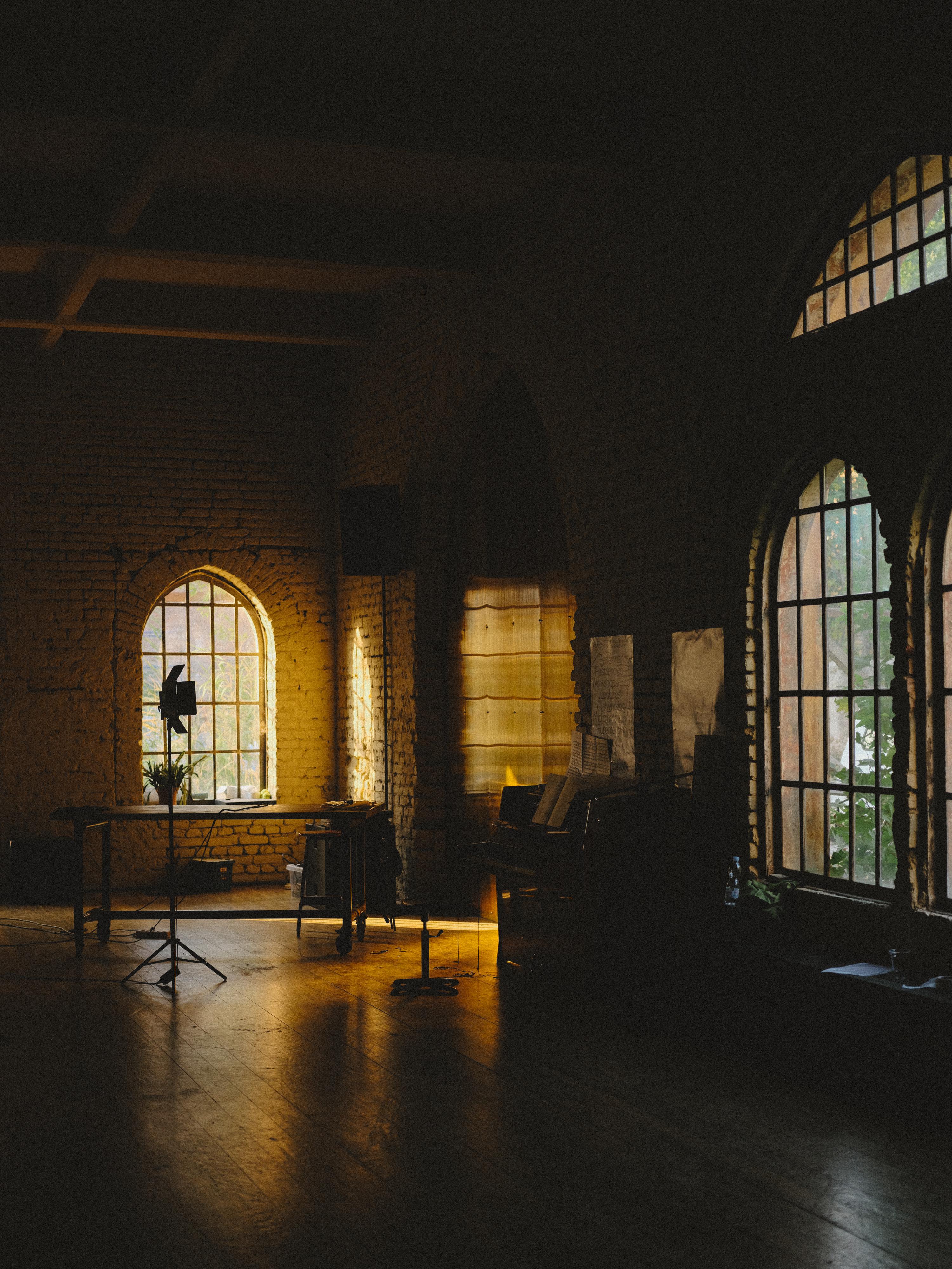 Industrial interior with brick walls, large arched windows, wooden floors. Warm light illuminates workspace with easel and equipment.