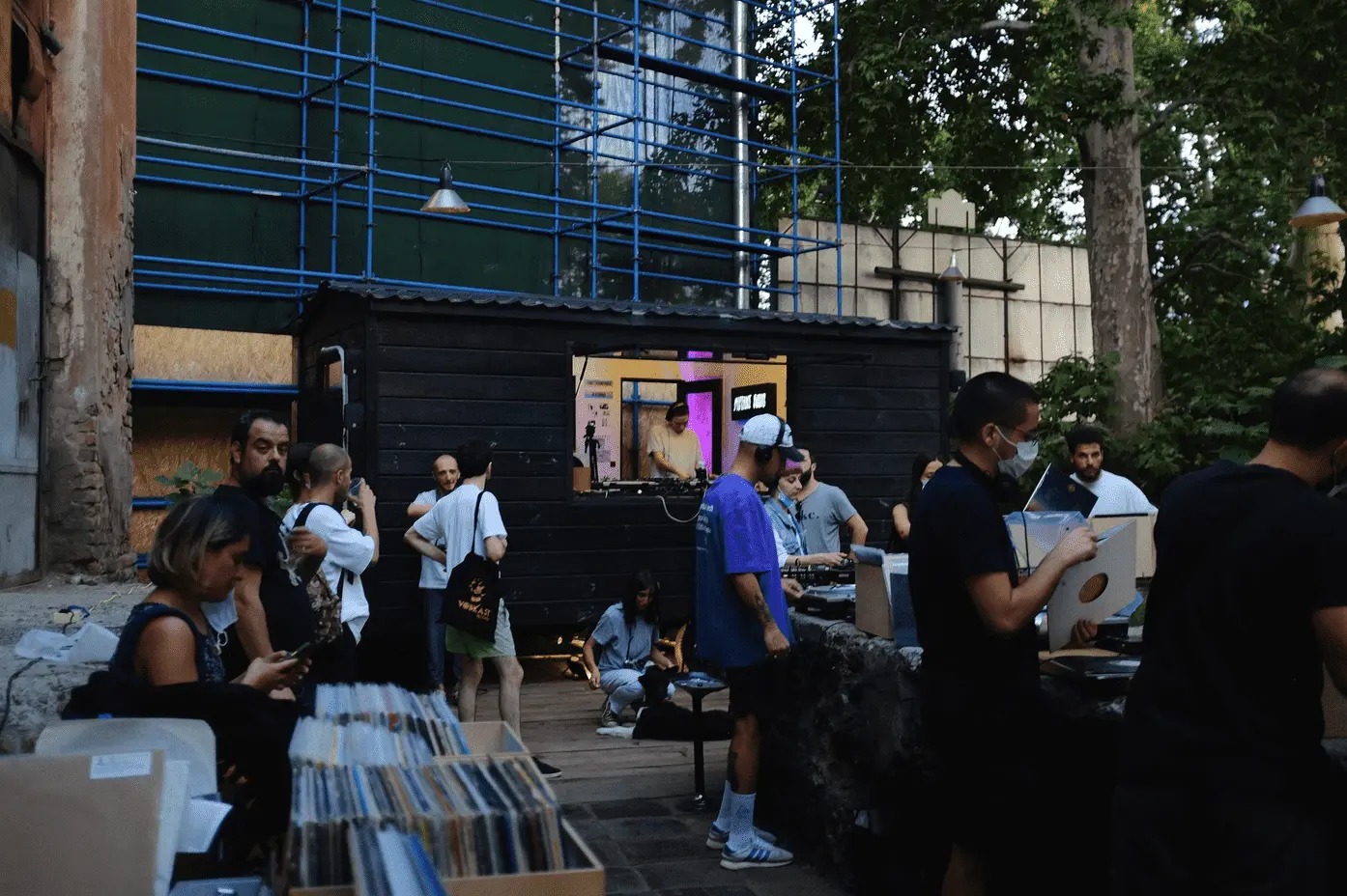 People browsing records at outdoor market stall beneath industrial building with green scaffolding and trees overhead.