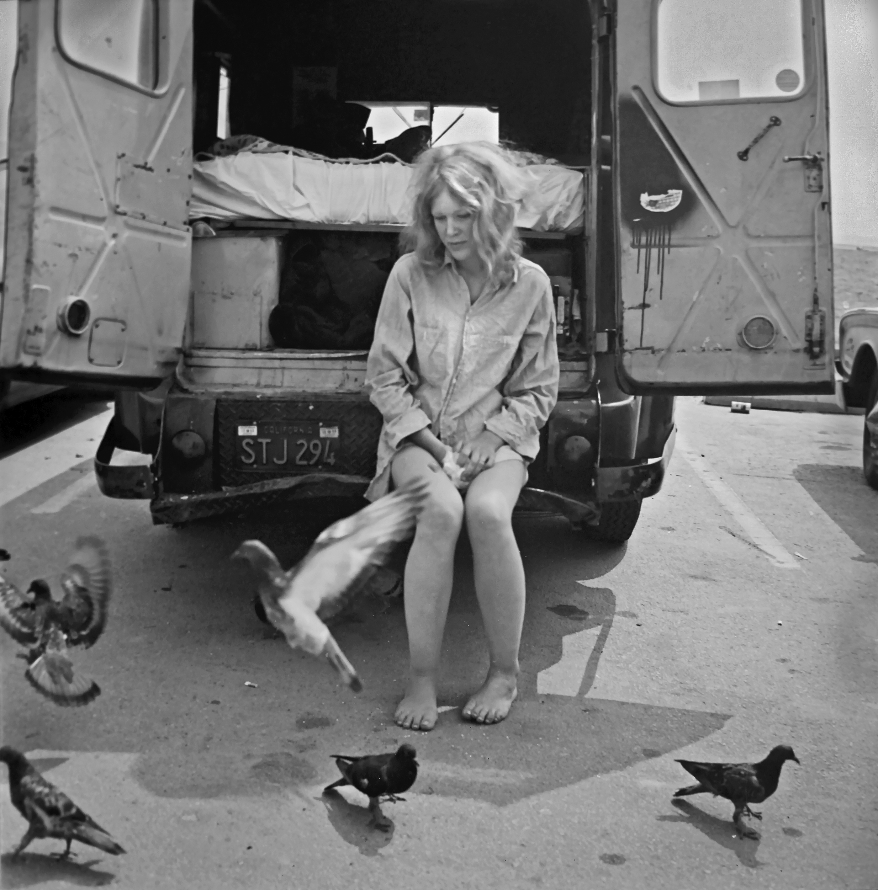 A black and white image showing a person, likely a woman, sitting on the back of a vehicle surrounded by pigeons on the ground.