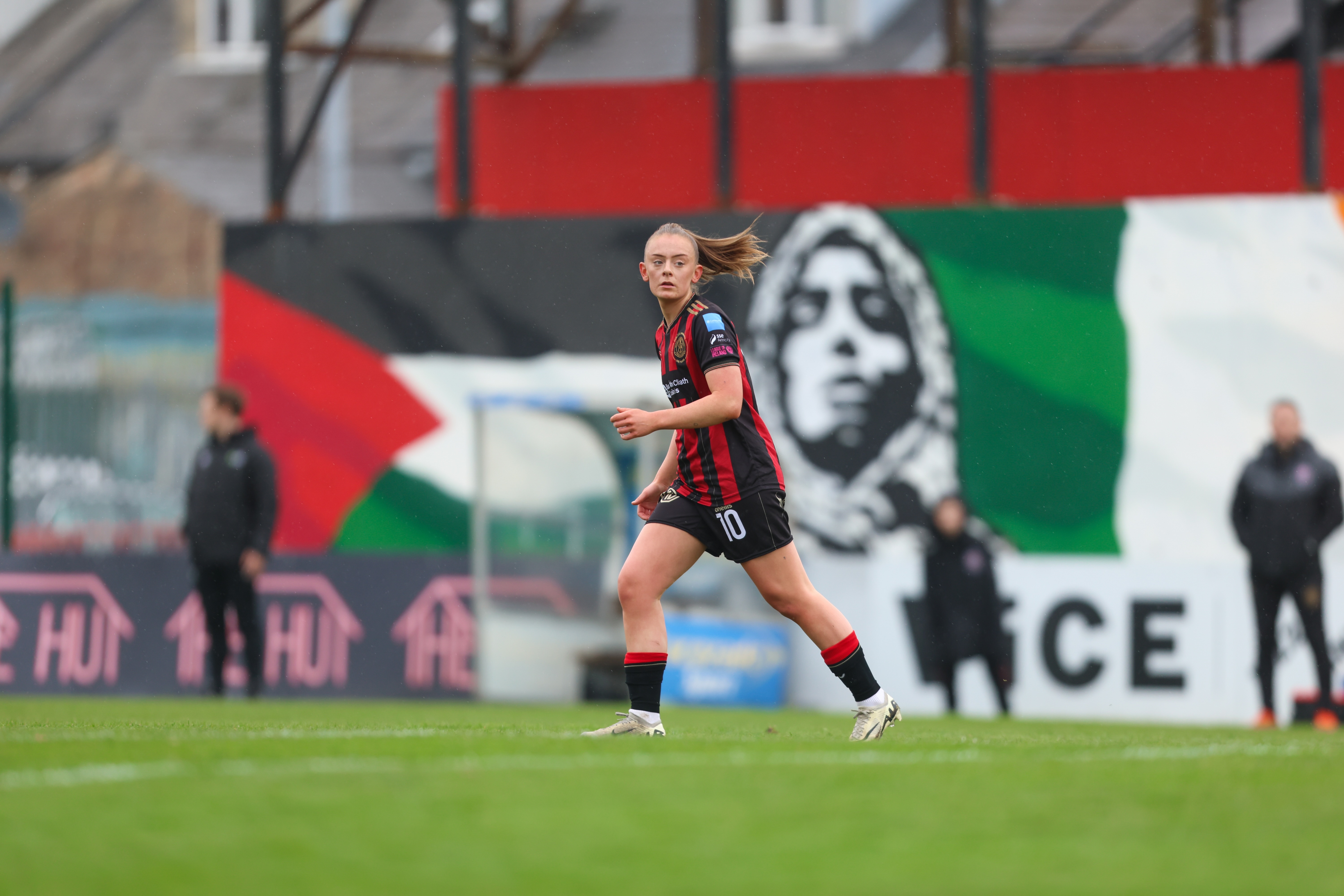 A woman in a red football jersey, shorts and cleats running on a pitch in front of a large, colourful banner.
