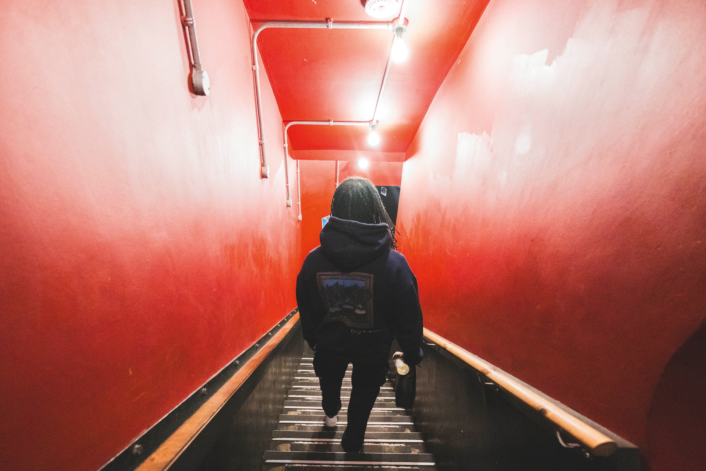 Person in dark clothing ascending wooden stairs in red-lit tunnel with handrails and overhead lighting fixtures.