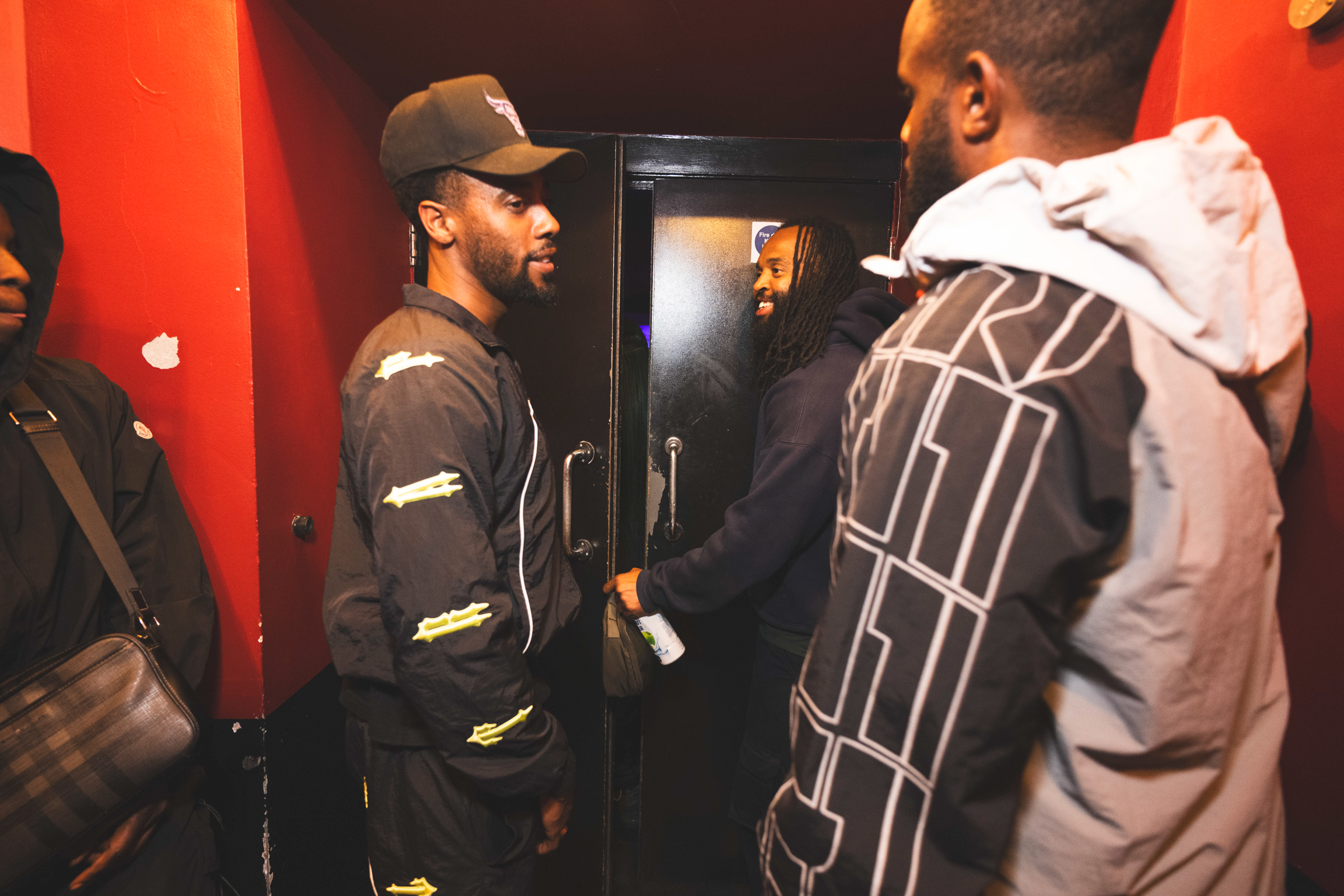Two men in conversation indoors - one wearing black cap and jacket, the other in grey hoodie with geometric pattern on back.