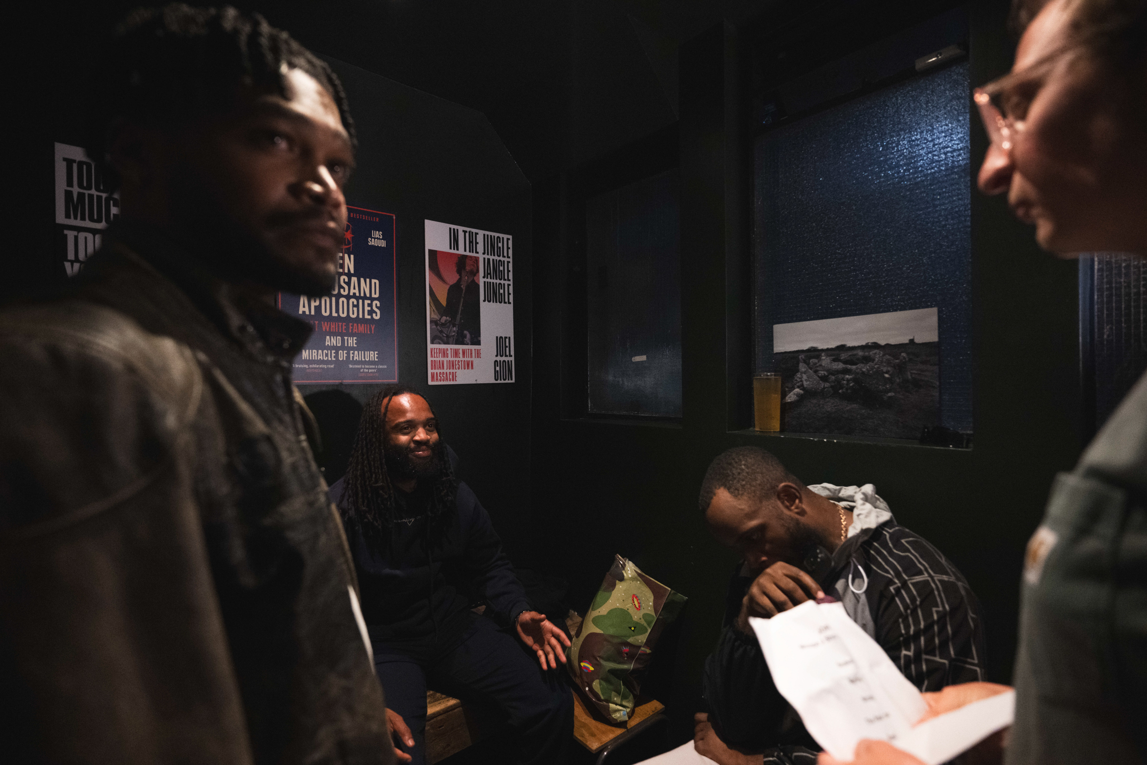 Two men in conversation in dimly lit room with posters on blue wall, third person seated in background holding papers.