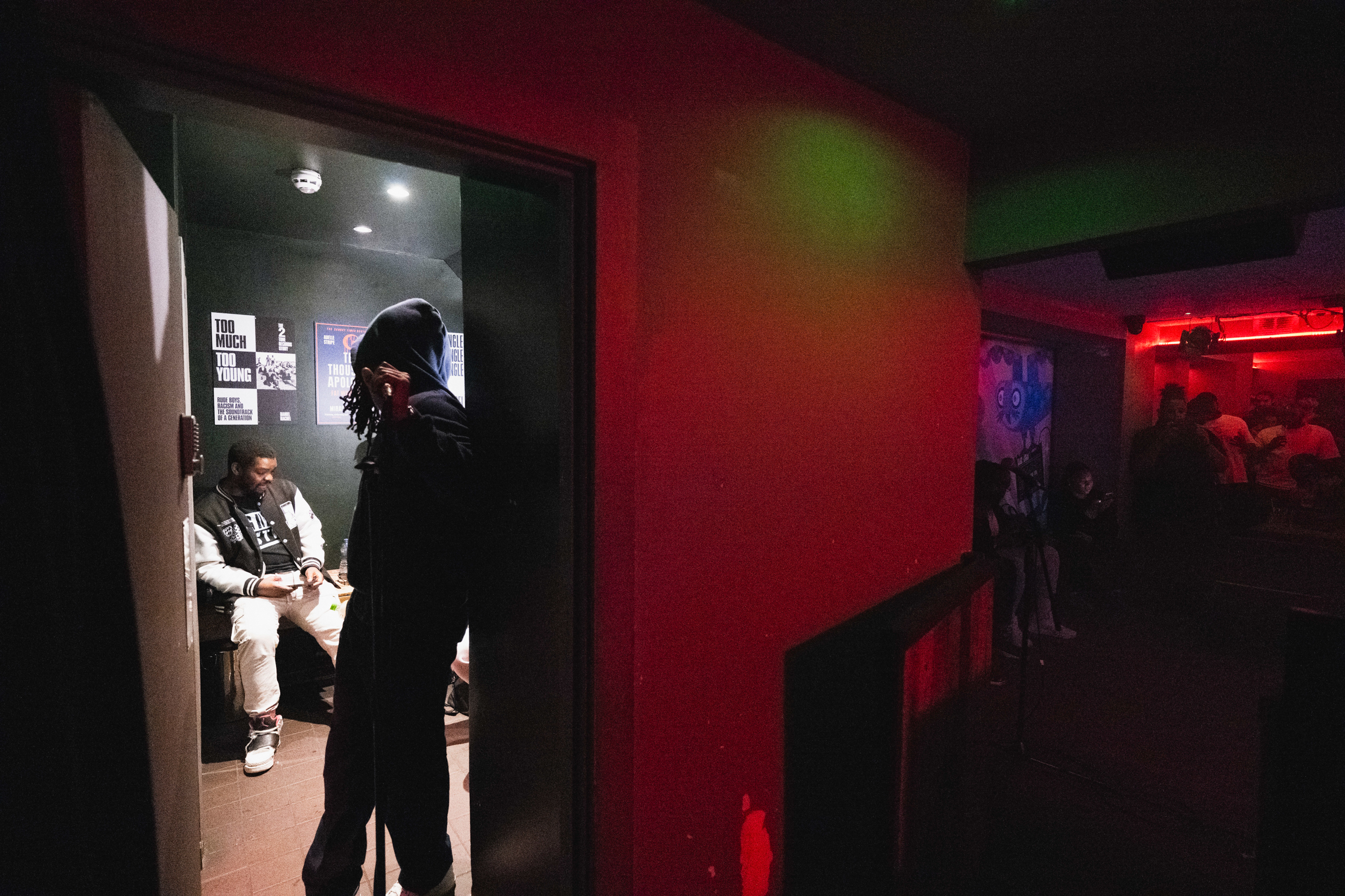 Dimly lit indoor space with vibrant red and green lighting. Doorway on left shows seated figure, silhouettes visible on right side.