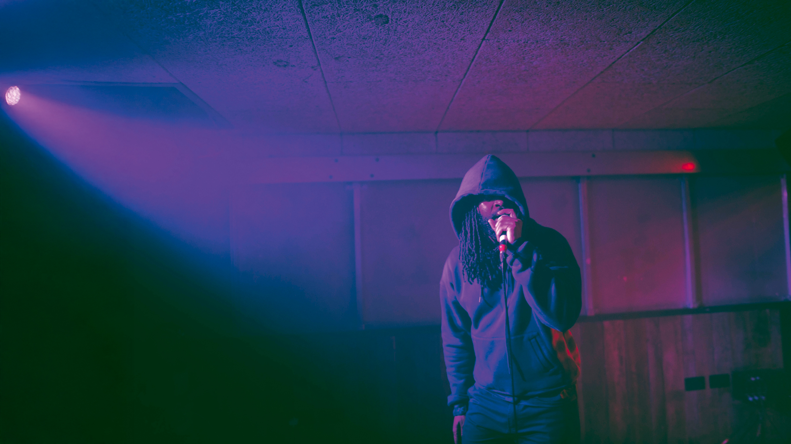 Person in hooded jacket holding microphone in dimly lit room with purple and blue lighting, industrial ceiling visible above.