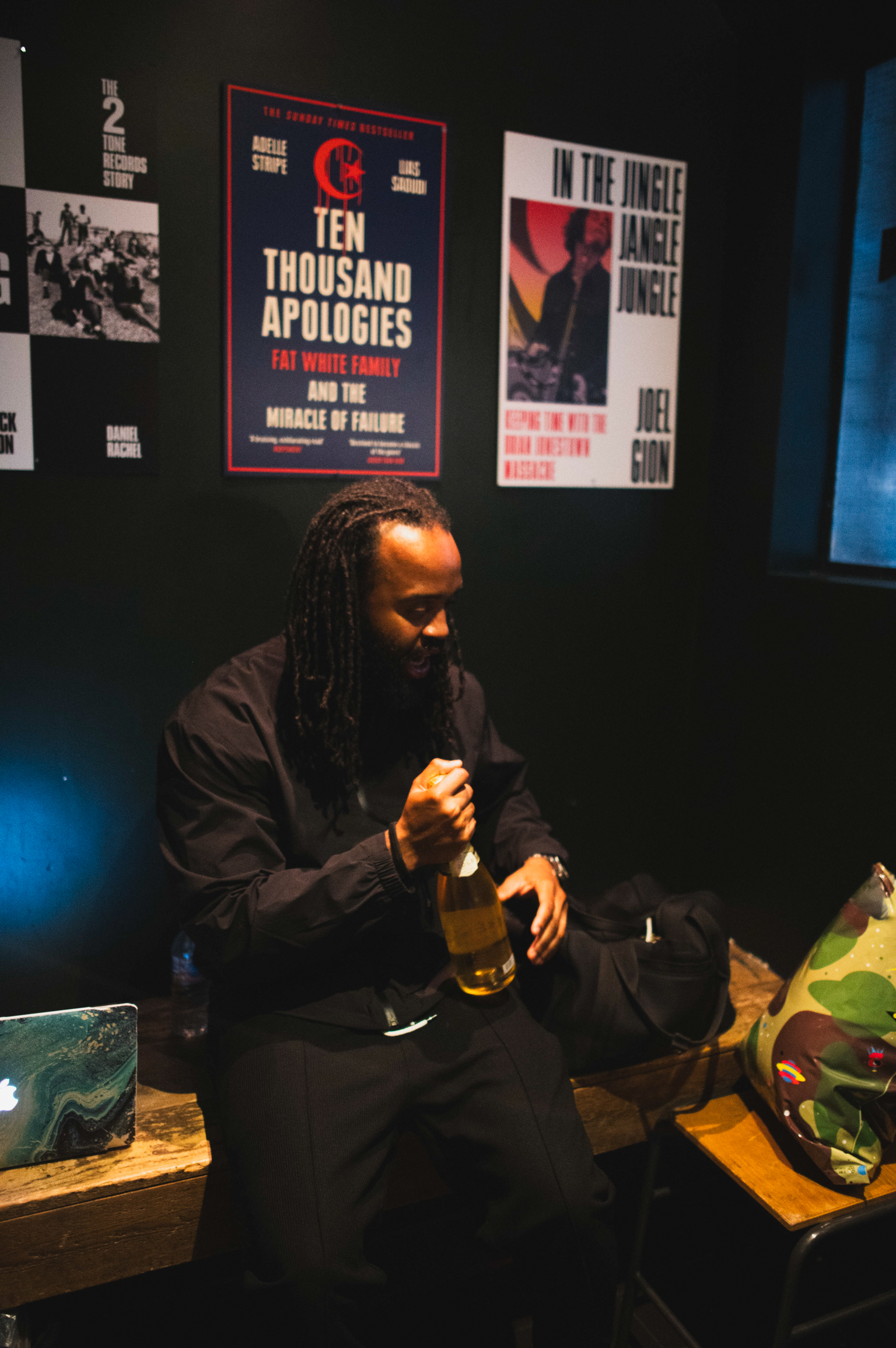 Man with dreadlocks sits at desk with laptop in dimly lit room. Book posters on dark wall behind, including "Ten Thousand Apologies".