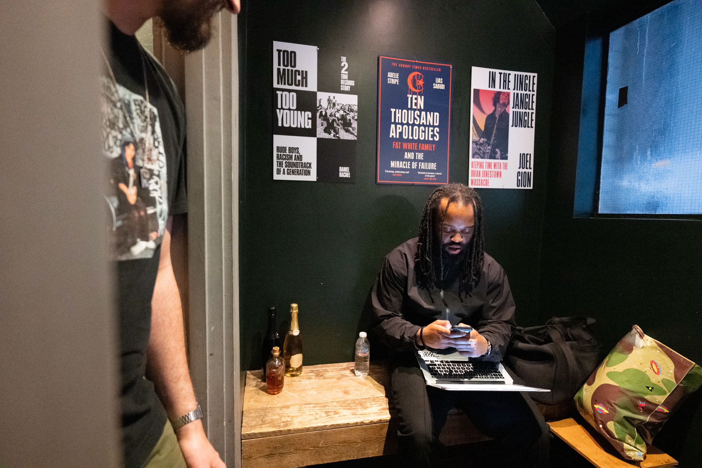 Man with headphones at laptop in dark room with posters on wall, bottles on wooden table, blue screen visible right side.