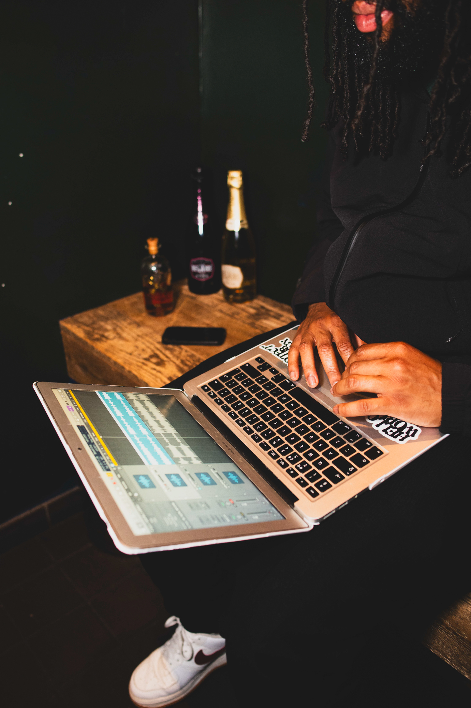 Person typing on open laptop displaying software interface, wooden table with bottles and items, dimly lit room with warm lighting.