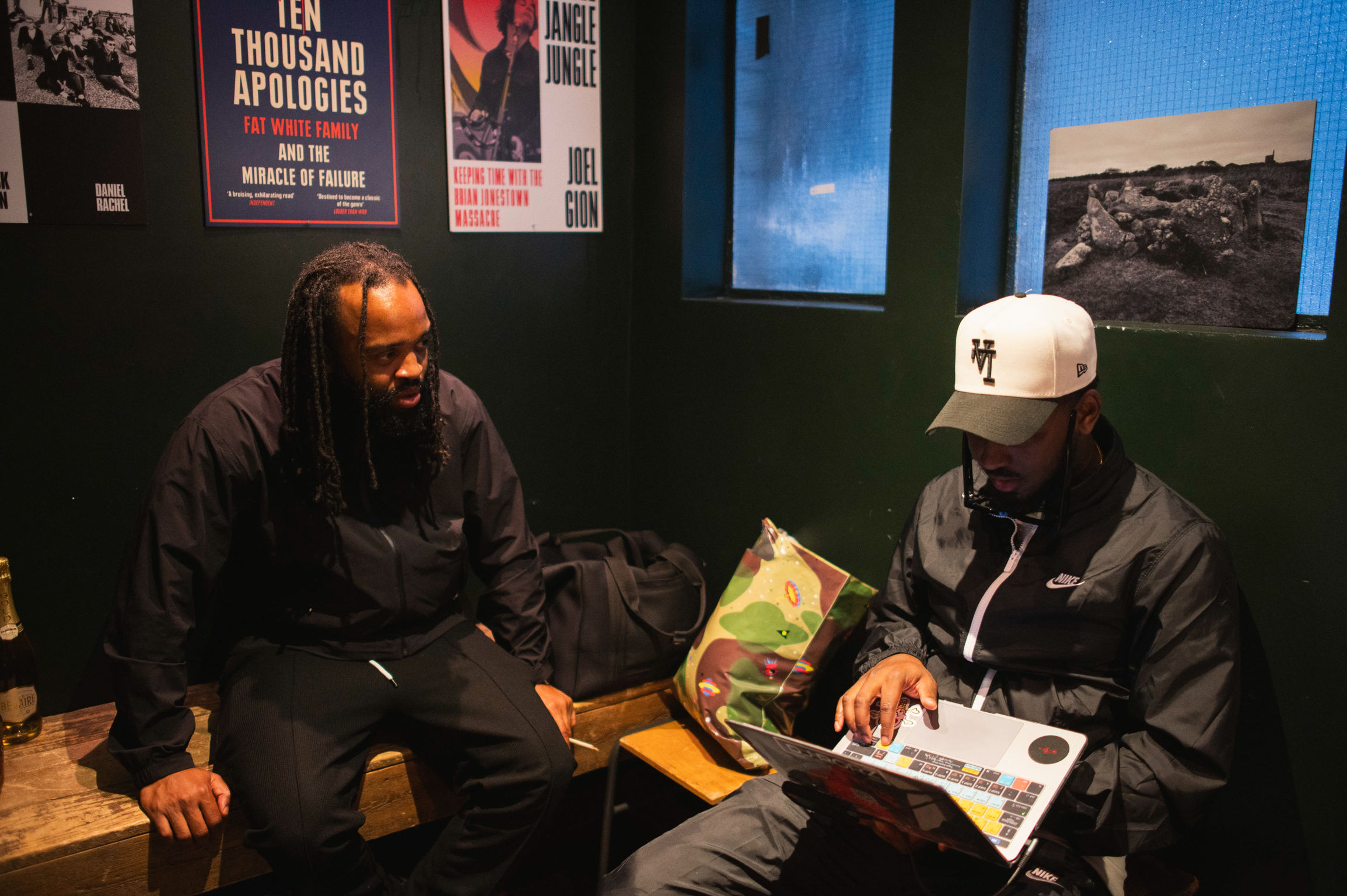 Two men sitting in room with dark walls, one wearing headphones, other in white cap reading magazine. Posters and framed images on walls.