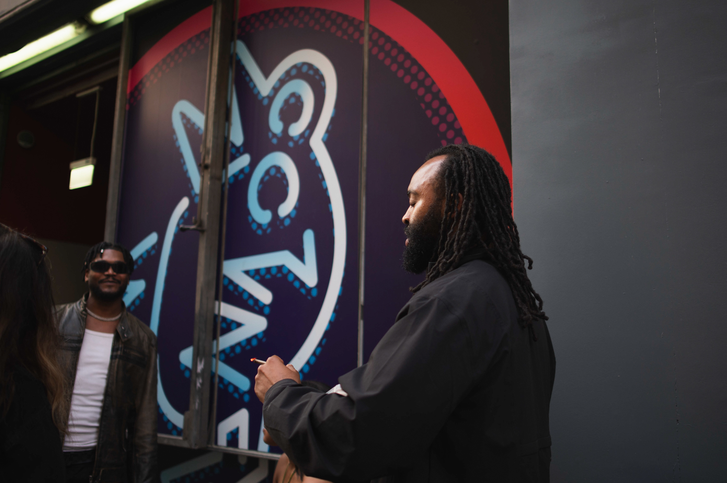 Man with dreadlocks painting blue and white graffiti design on wall whilst another person watches in background.