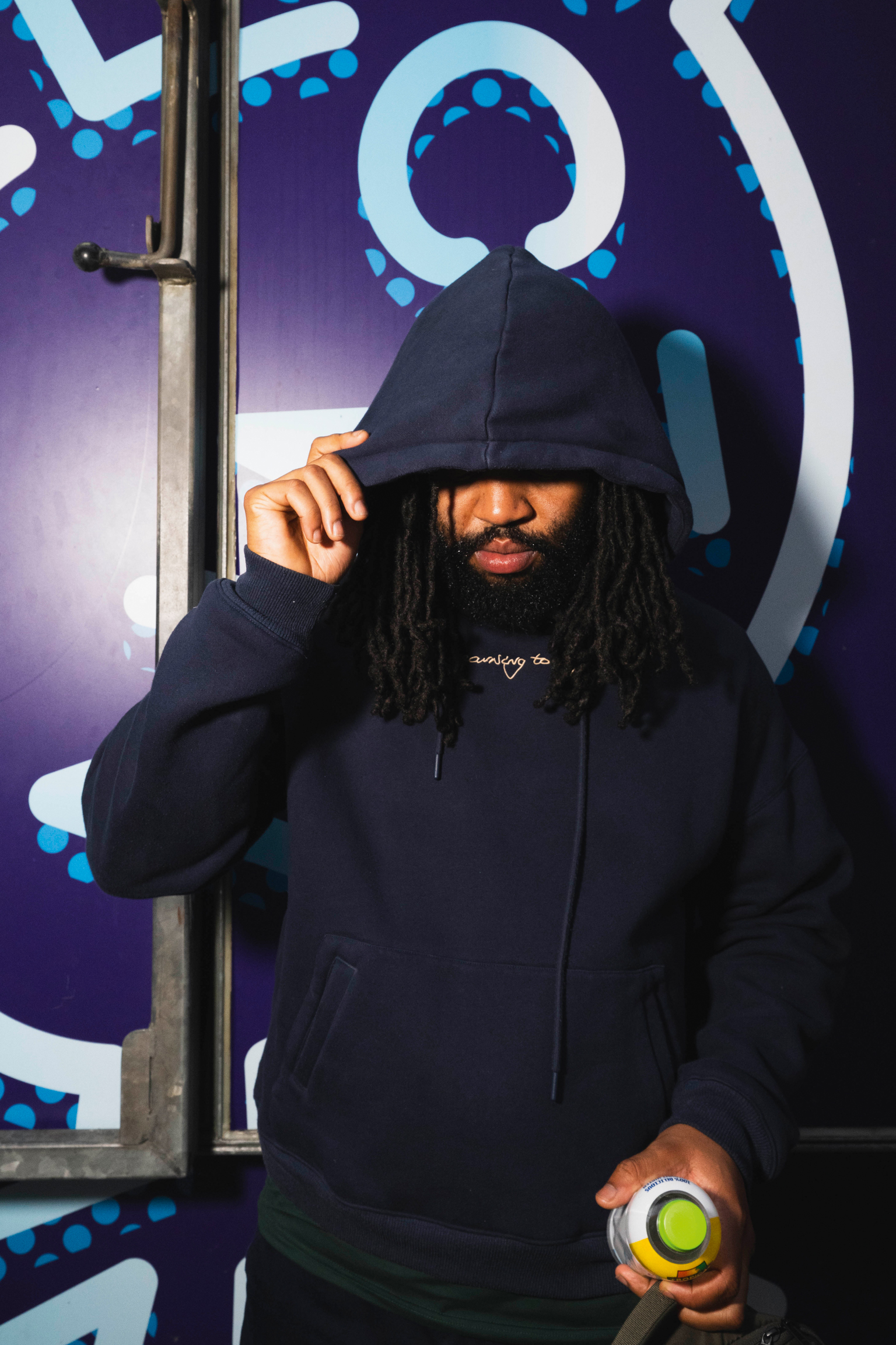 Person in black hoodie with locs holding green circular object, standing against purple wall with blue and white geometric patterns.