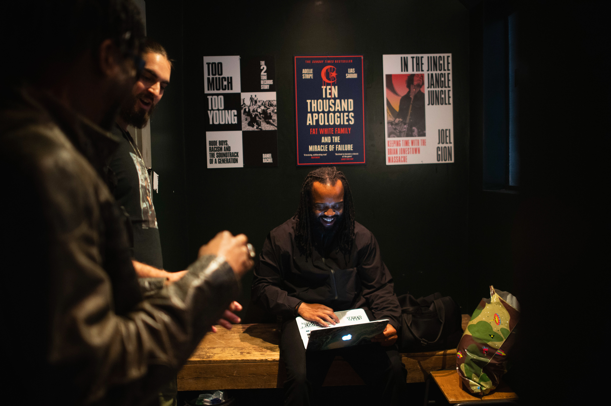 Two men in dark interior with book posters on wall behind them, one standing, one seated reading, warm lighting on wooden surface.