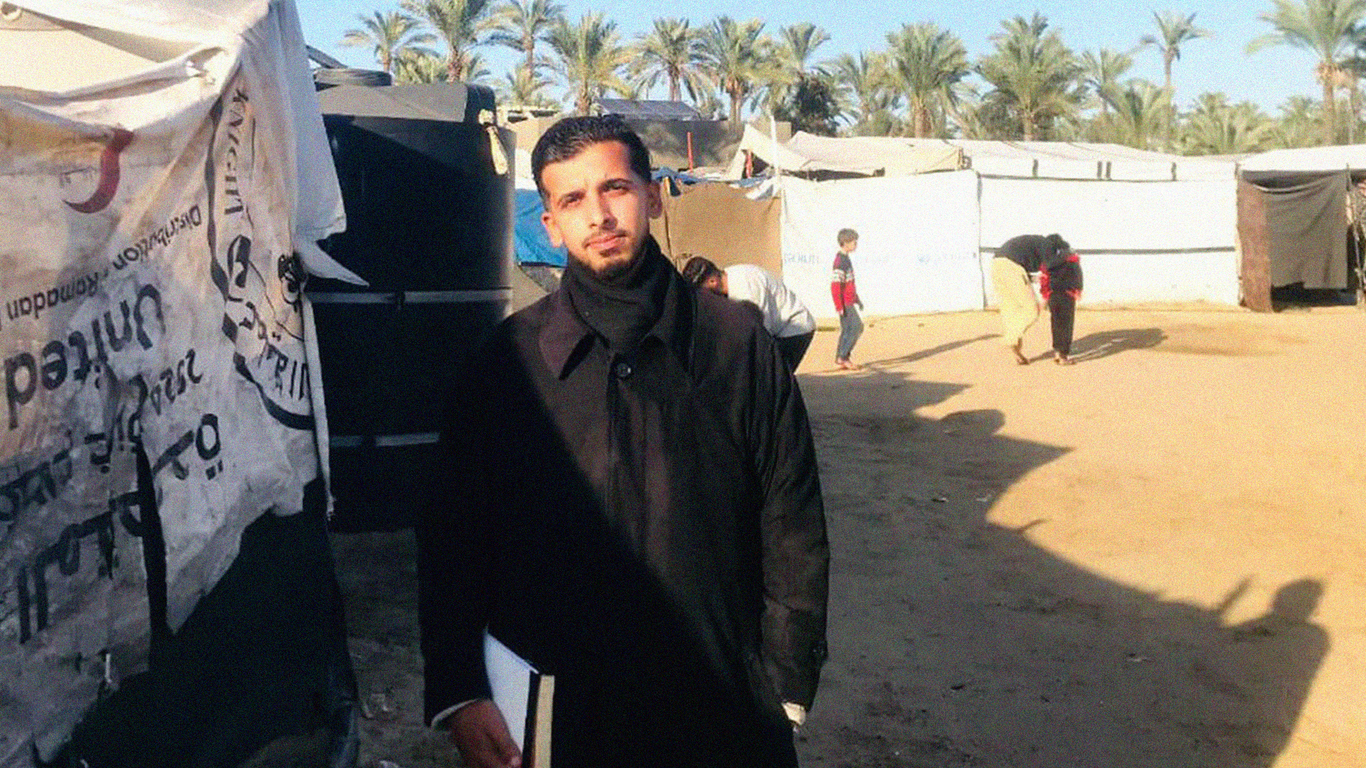 Man in dark jacket standing beside white tents in sandy area with palm trees and buildings in background under clear sky.