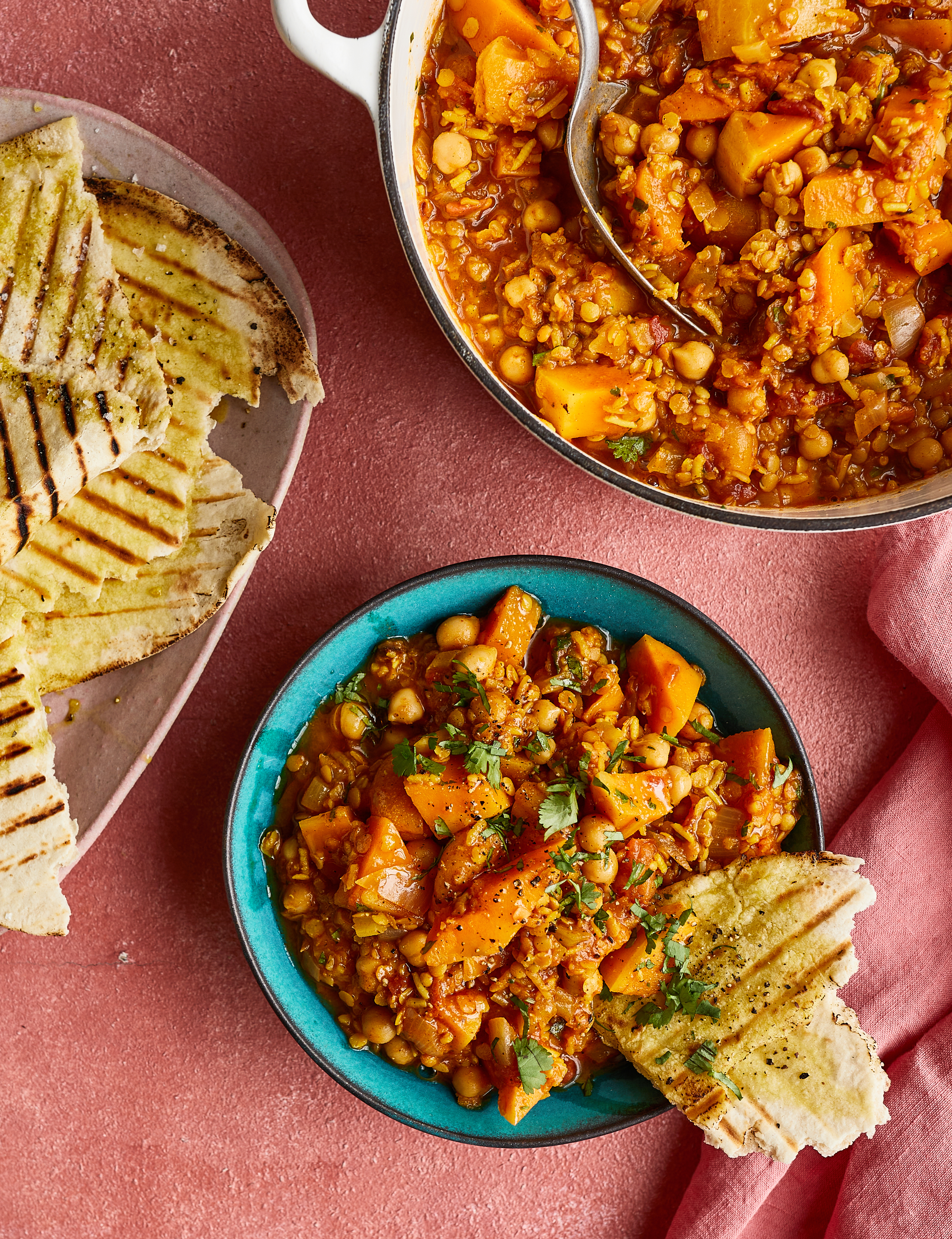 Lentil curry with sweet potato in blue bowl and steel pan, served with grilled naan bread on terracotta background.