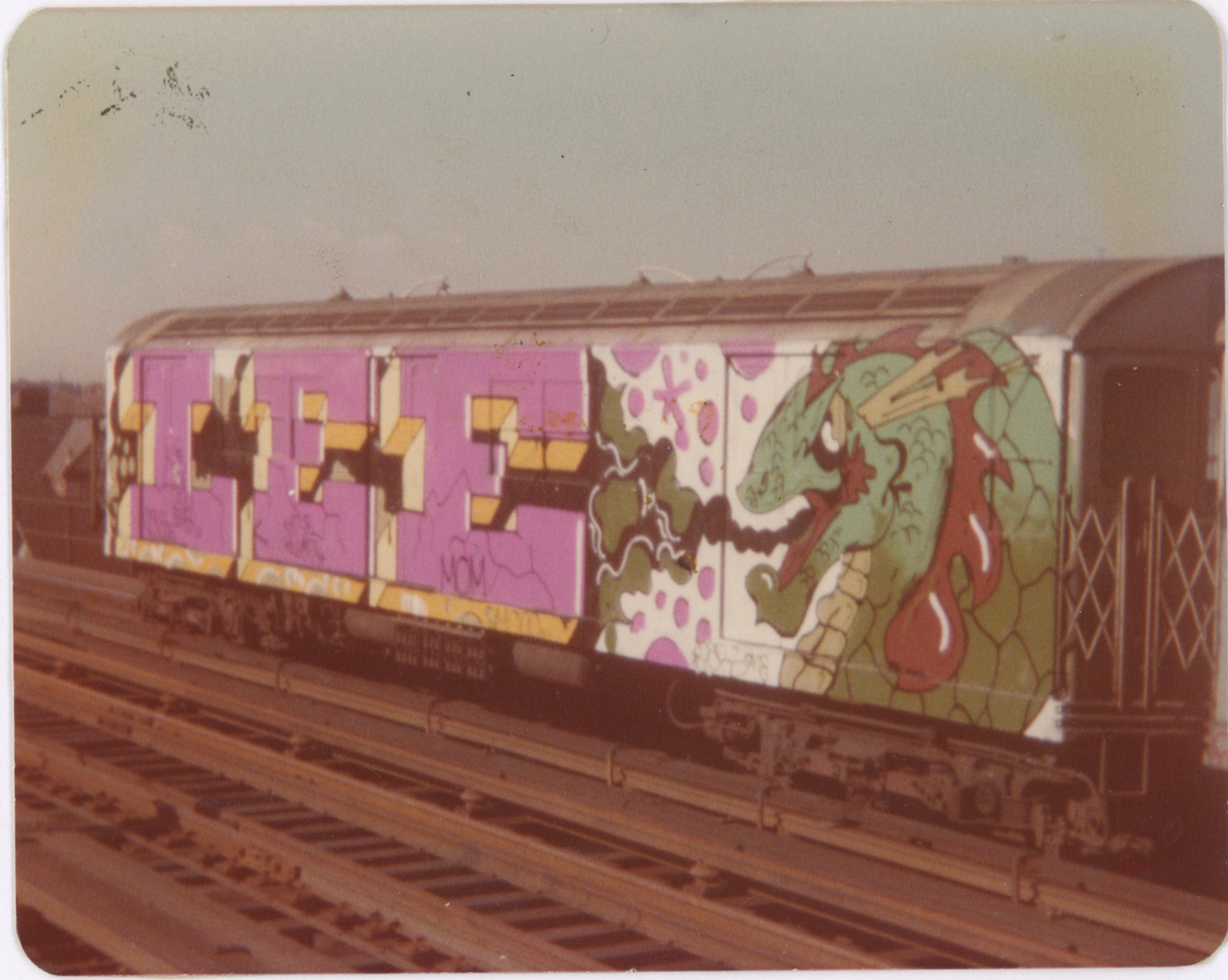 Train carriage with graffiti featuring purple and yellow block letters, green dragon design, and colourful artwork on railway tracks.