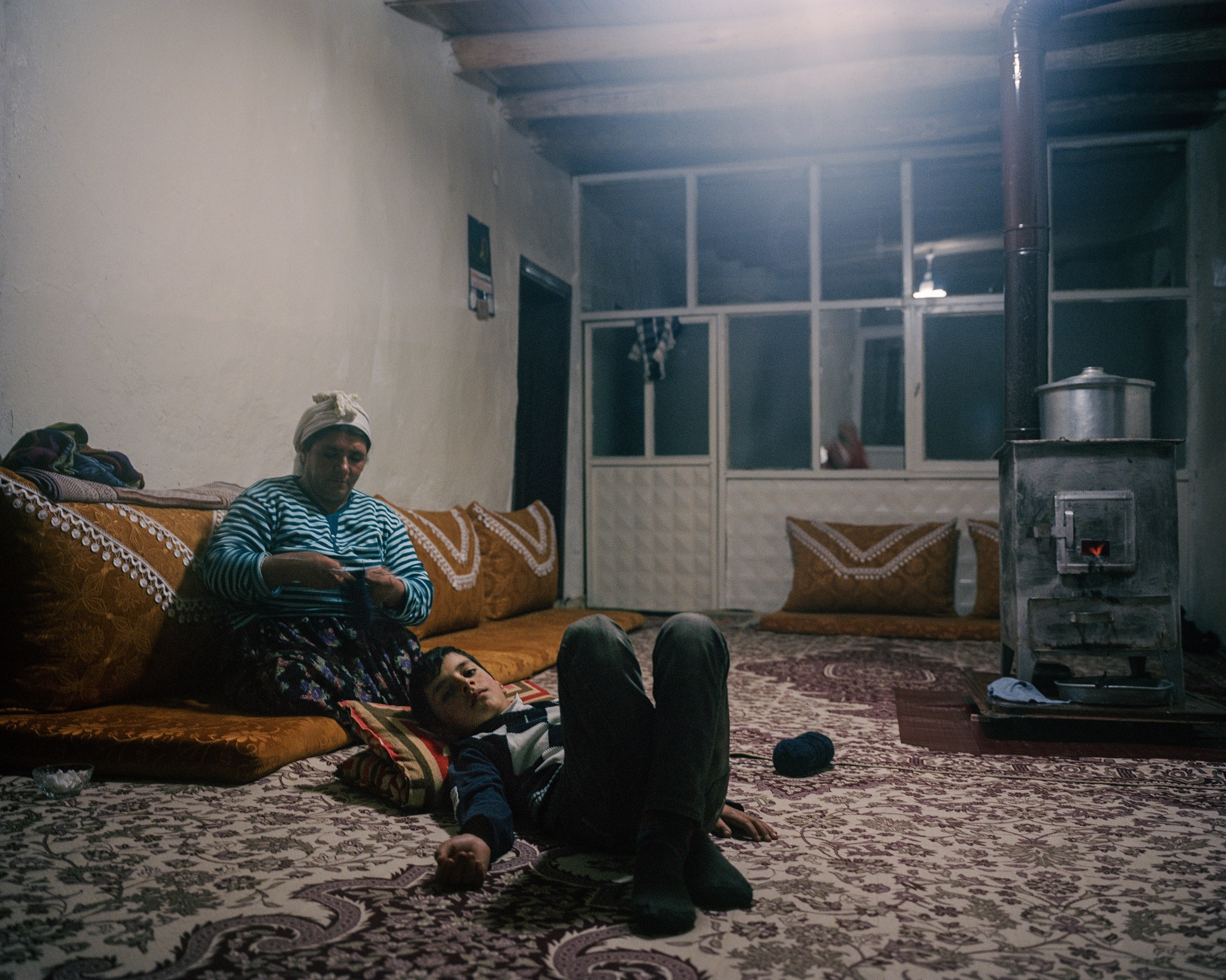 Cosy room with woman seated on sofa and child lying on floor, both wrapped in blankets near a wood-burning stove.