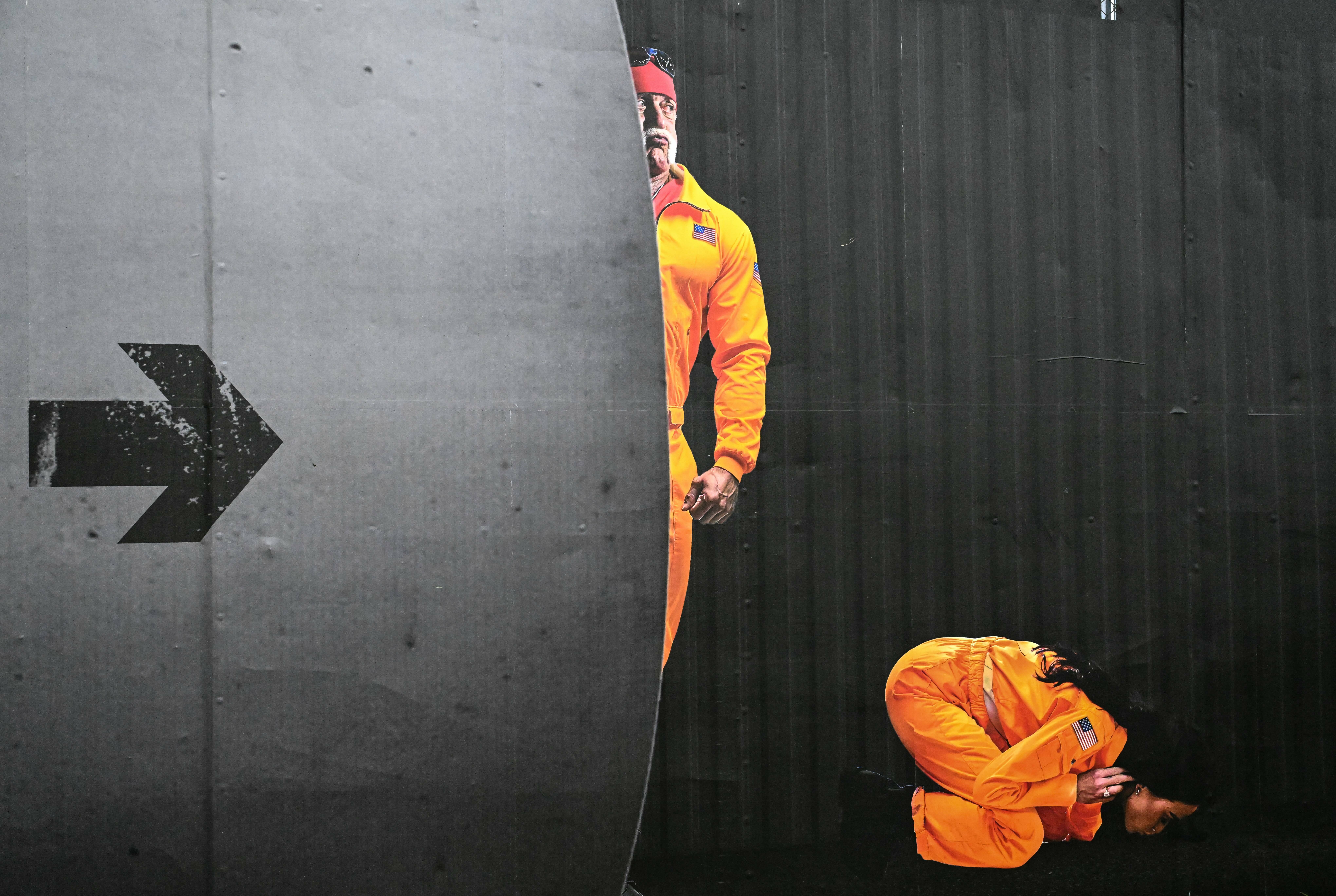 Two figures in bright orange jumpsuits against grey concrete wall with black arrow pointing right. One figure hanging from wall, another crouched below.