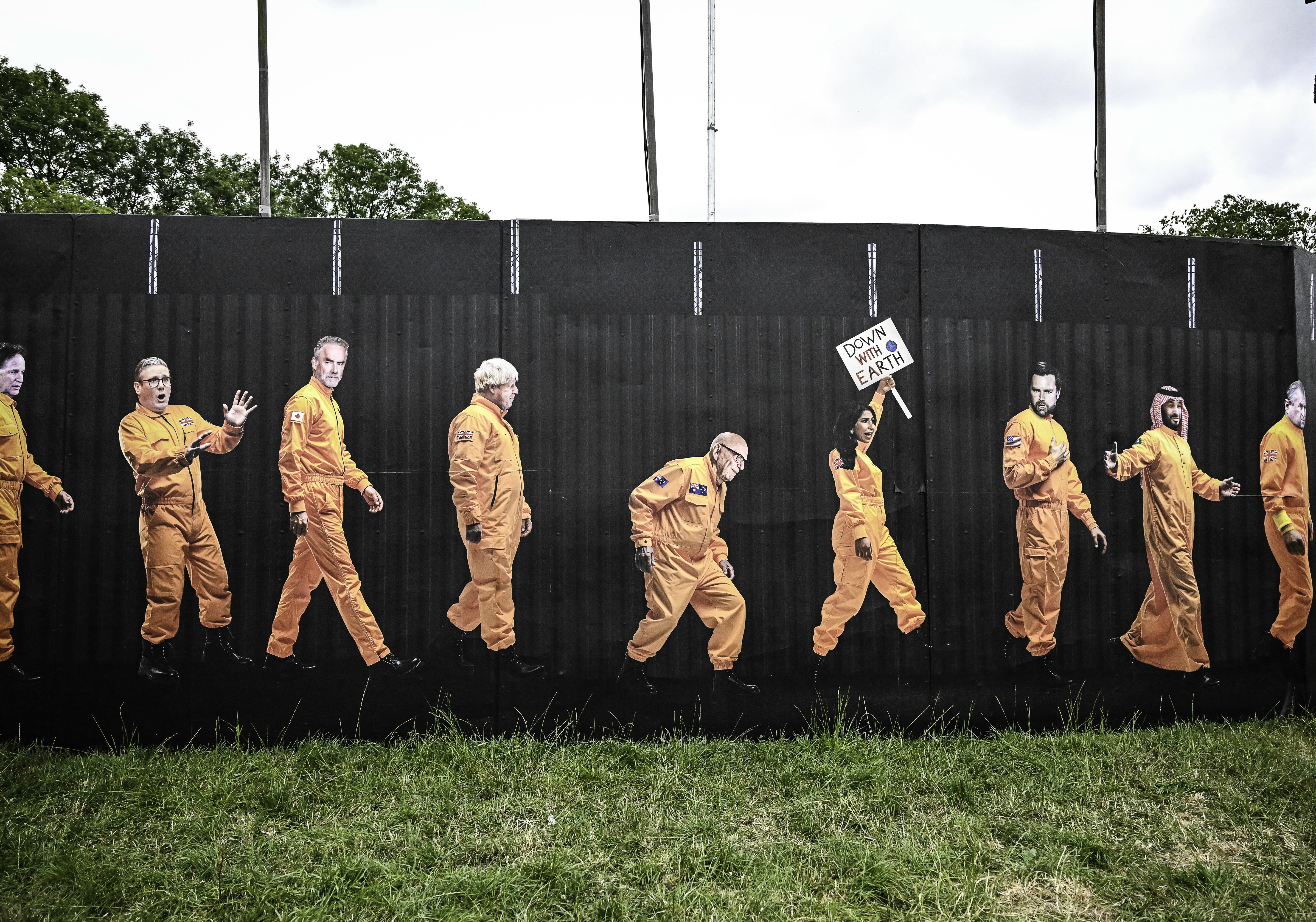 Street art mural on black fence showing figures in orange prison uniforms walking in line, one holding a sign, with green grass below.