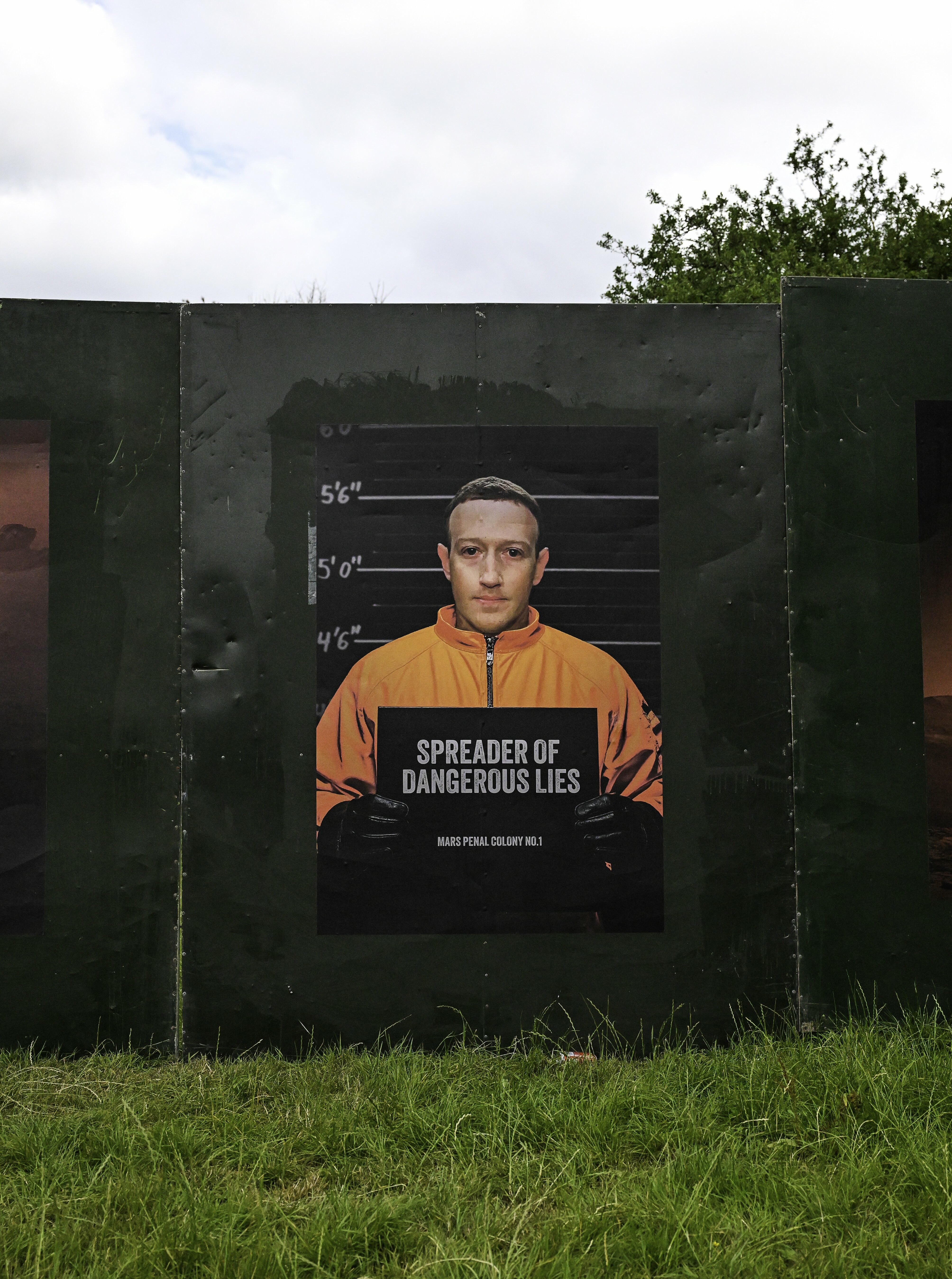 Street art mural showing mugshot-style portrait of man in orange jumpsuit holding sign reading "SPREADER OF DANGEROUS LIES" on green wall.