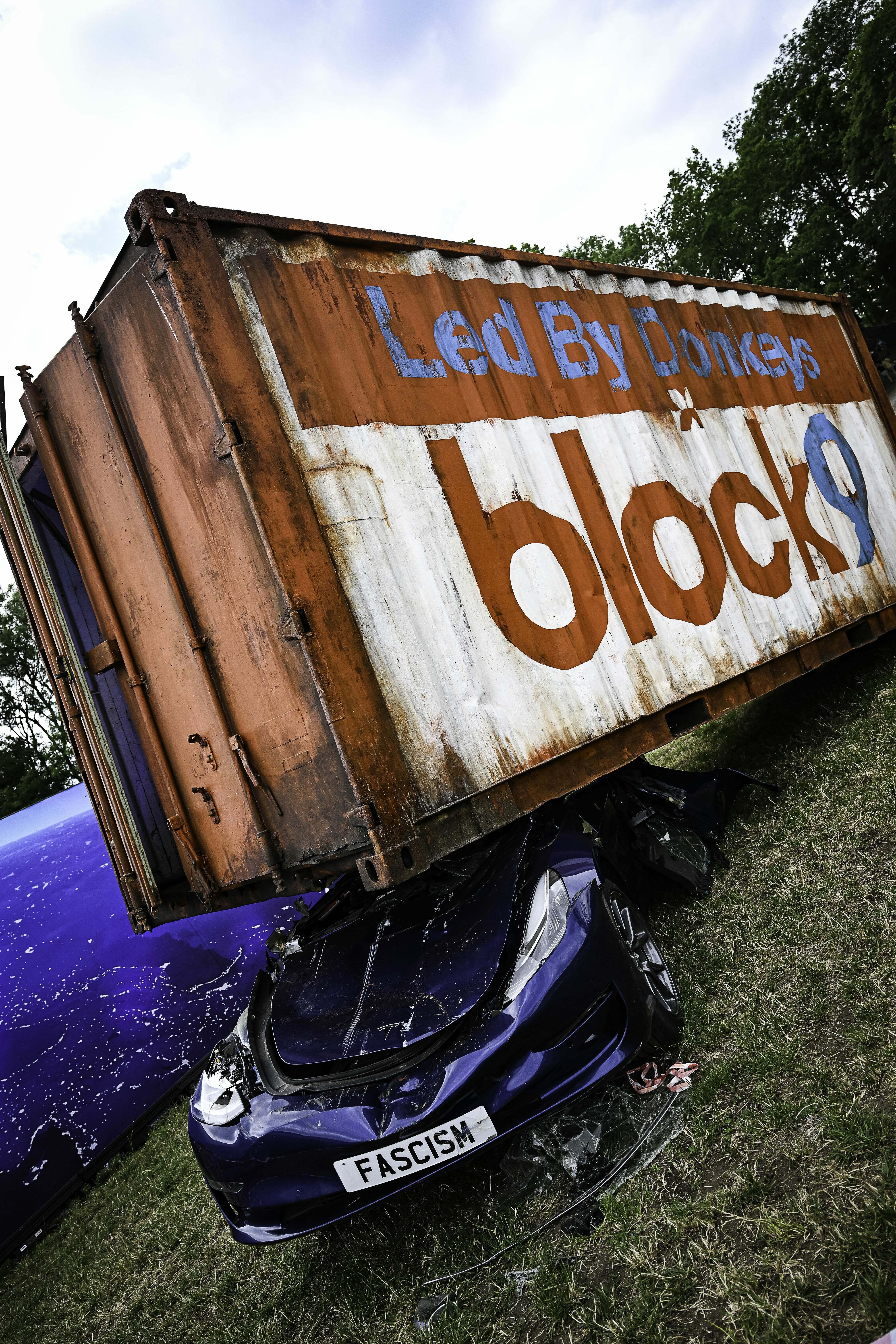 Rusty orange shipping container with "Led By Donkeys" and "block?" painted in blue and white text, crushing blue car below.
