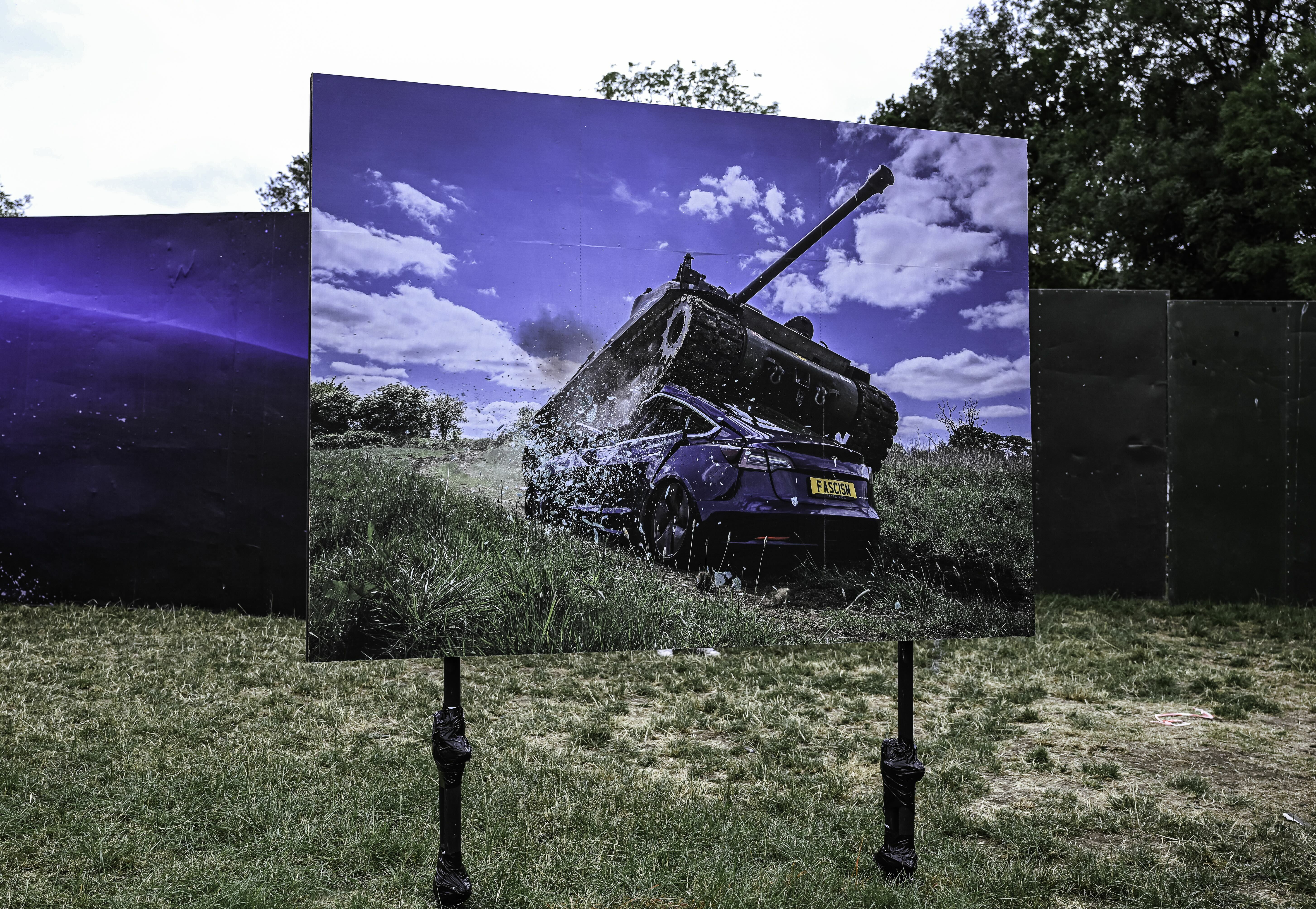 Large outdoor display panel showing military tank in green field under blue sky with white clouds, flanked by darker panels.