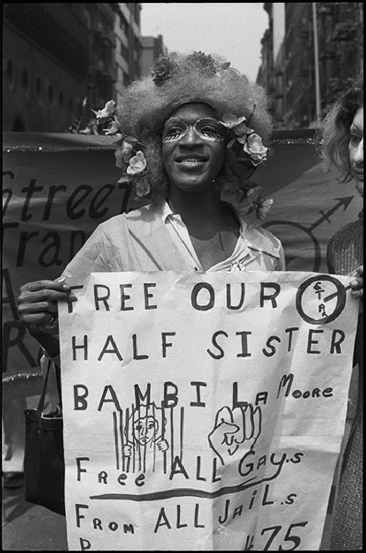 Black and white image of a person wearing a flower headpiece holding a sign that reads "Free our half sister Bambill, Free all gays from all jails."
