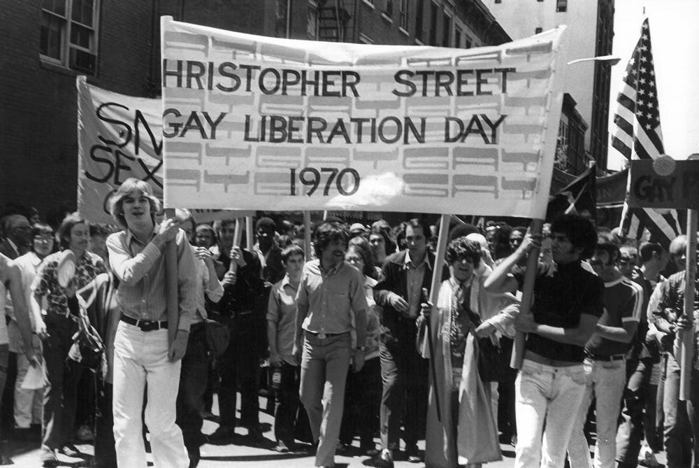 A large crowd gathered on a street, waving banners and flags during a pride parade event, captioned "Christopher Street Gay Liberation Day 1970".