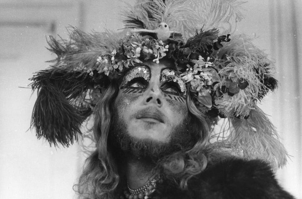 Woman's face adorned with intricate feathered crown, surrounded by flowers and foliage, black and white portrait.