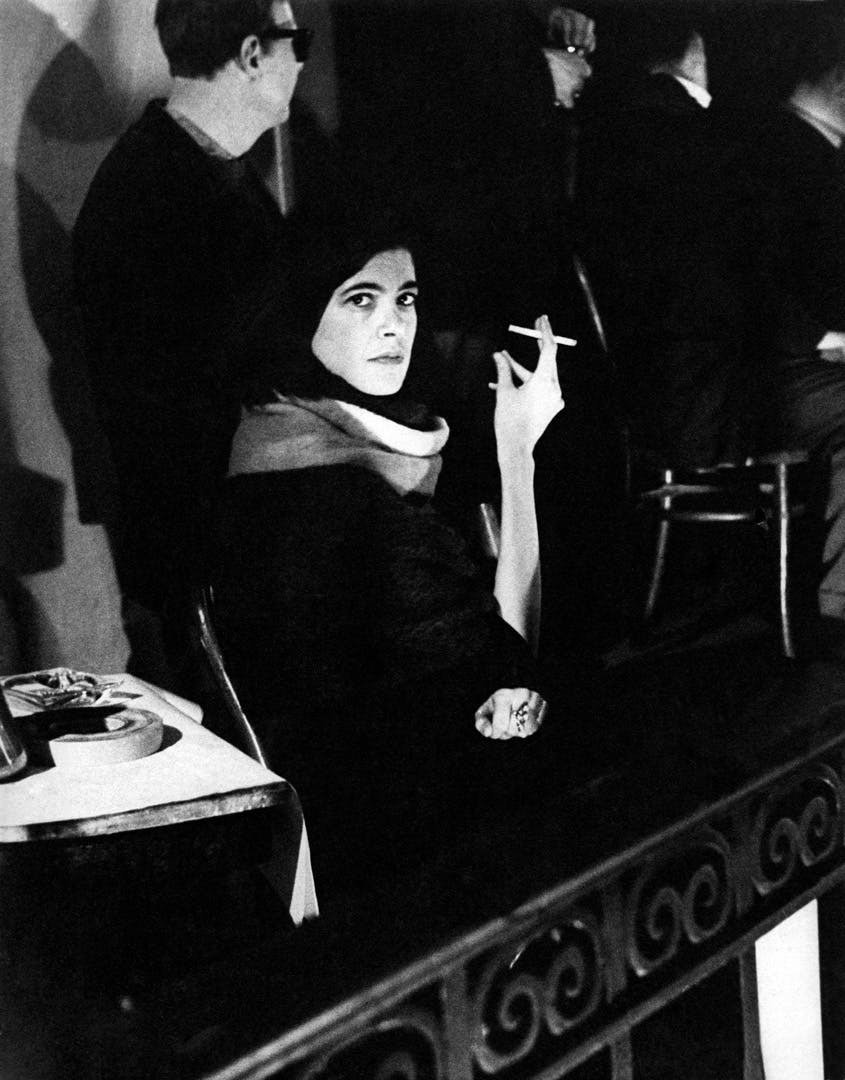 A black-and-white photograph showing a woman wearing a dark cloak, holding a cross, and standing on a staircase. Several other people, also in dark clothing, can be seen in the background.