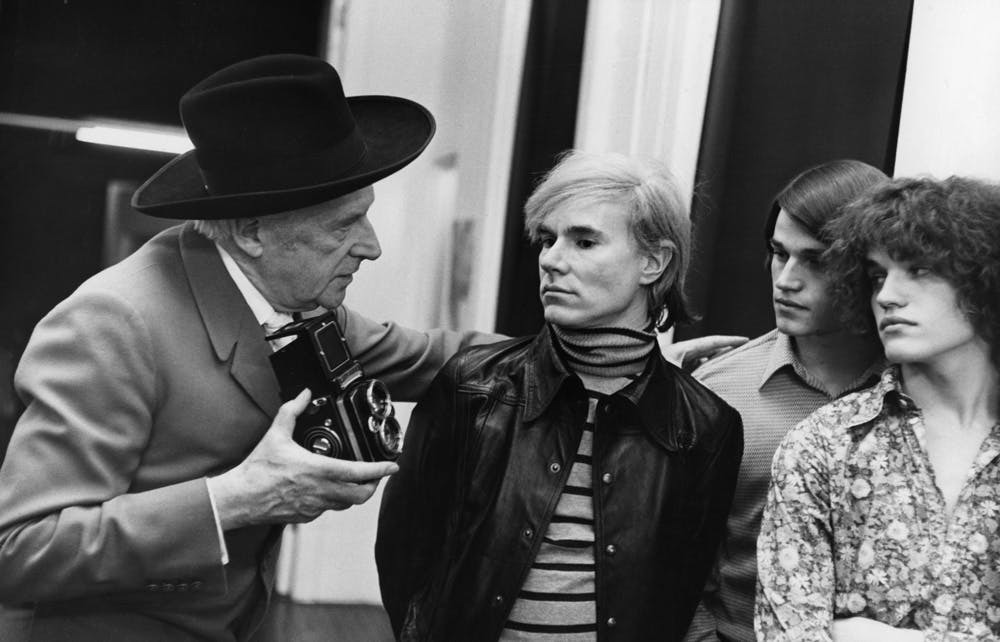 Four individuals, two men and two women, in black and white photograph. One man wears a hat and holds a camera, the other man and two women stand alongside him, all four wear casual attire.