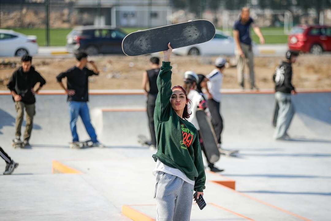Woman in green jumper holding skateboard in skate park, with other people and skaters in the background.
