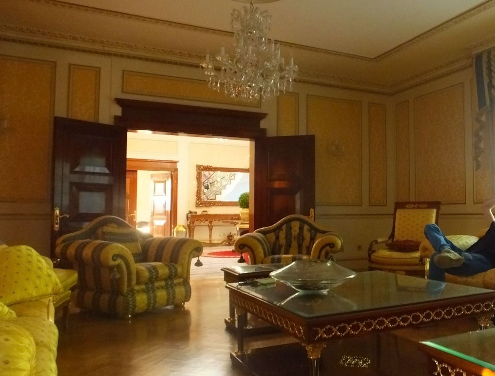 Elegant sitting room with crystal chandelier, striped sofas, ornate wooden coffee table, yellow walls with decorative moulding, and doorway to adjacent room.