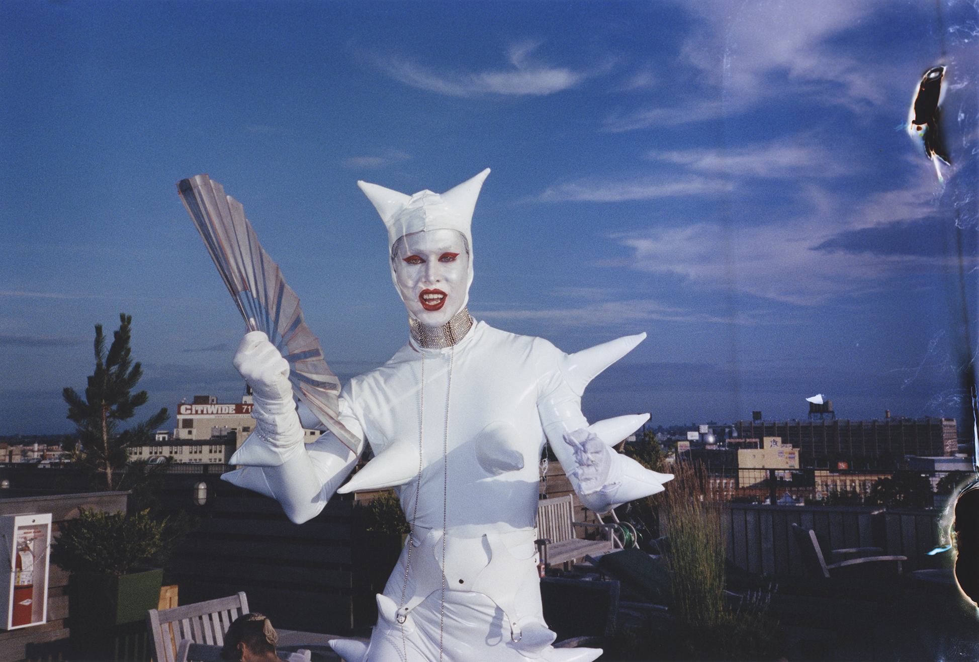 A person in an extravagant white costume with large, wing-like structures on their back and head, posing against a backdrop of a city skyline and blue sky with clouds.