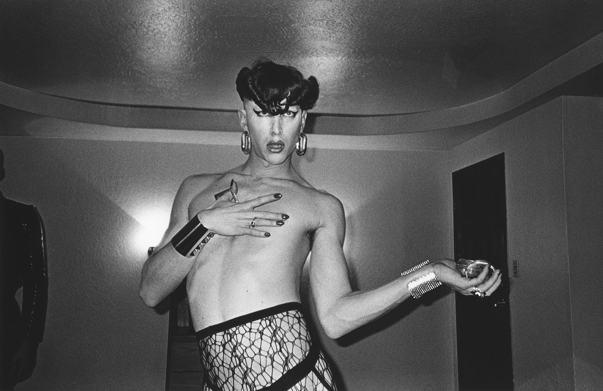 A black and white image shows a woman in a dramatic pose, her body partially obscured by her arms. She has an intense expression on her face and elaborate hairstyle.
