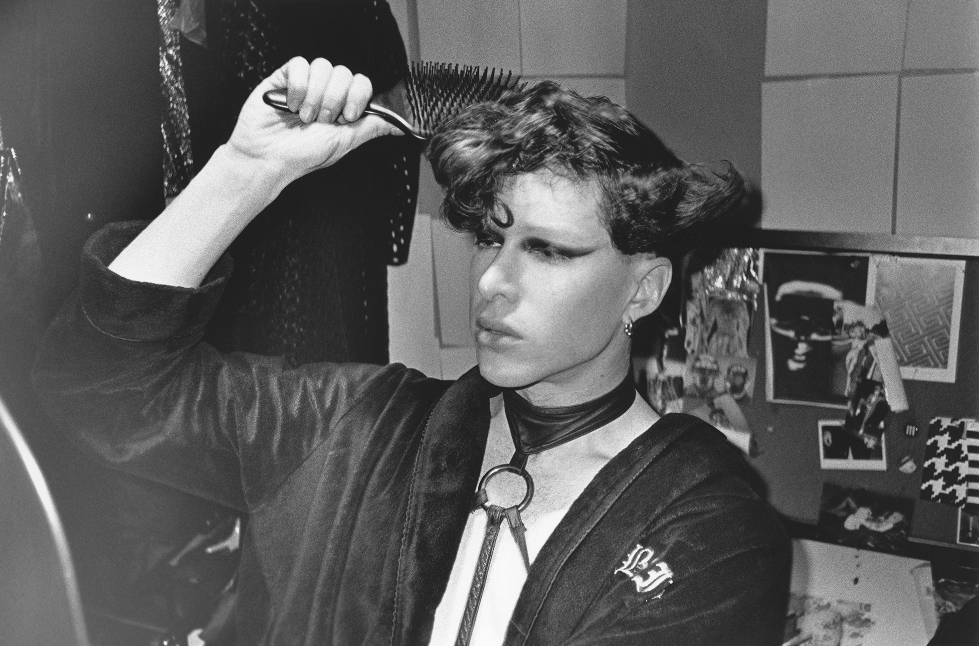 Monochrome photograph of a person brushing their curly hair and looking directly at the camera with a slightly stern expression. The photograph appears to have been taken in a domestic setting, with shelves visible in the background.