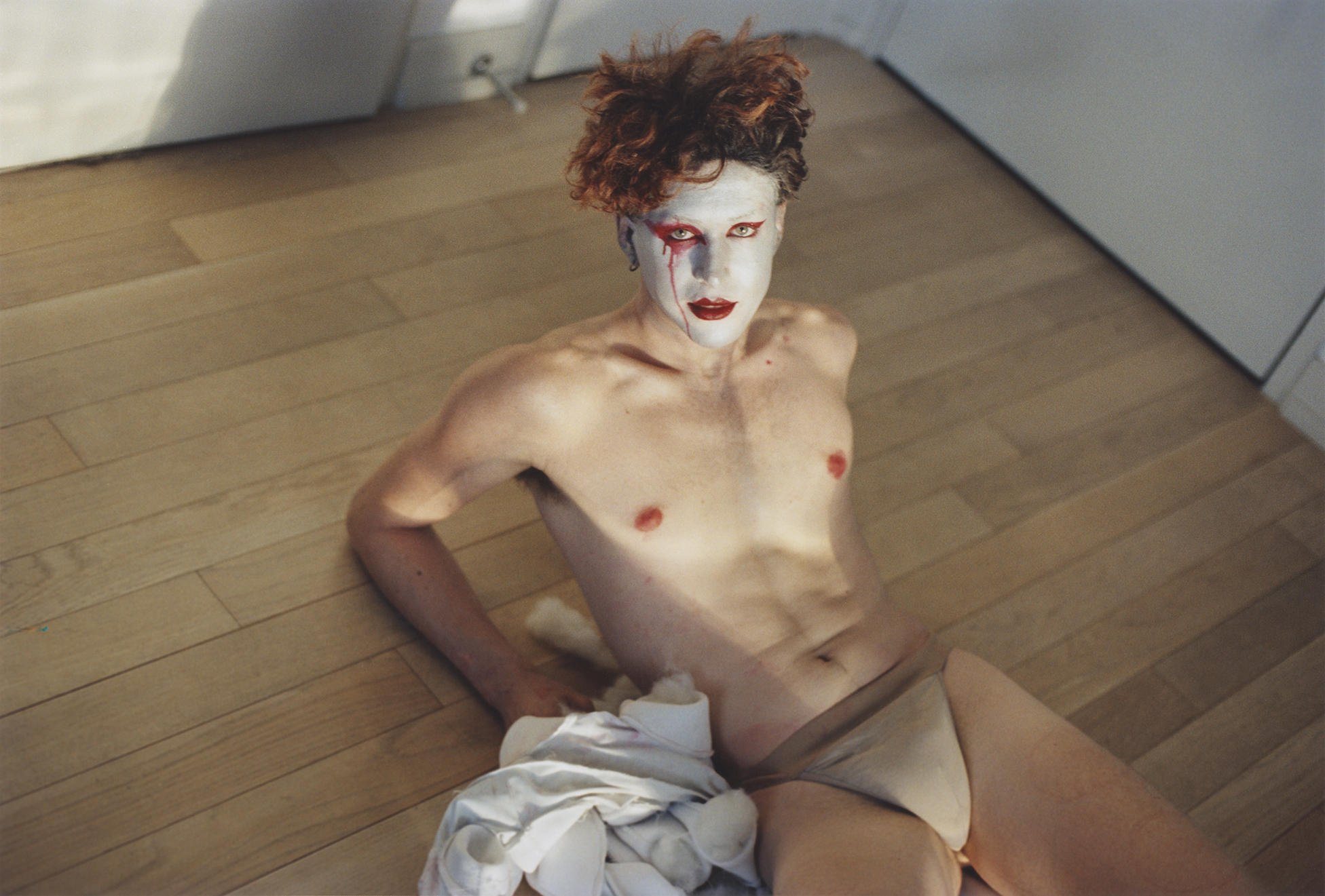 Shirtless figure with tousled red hair, white makeup and body paint, sitting on the floor.
