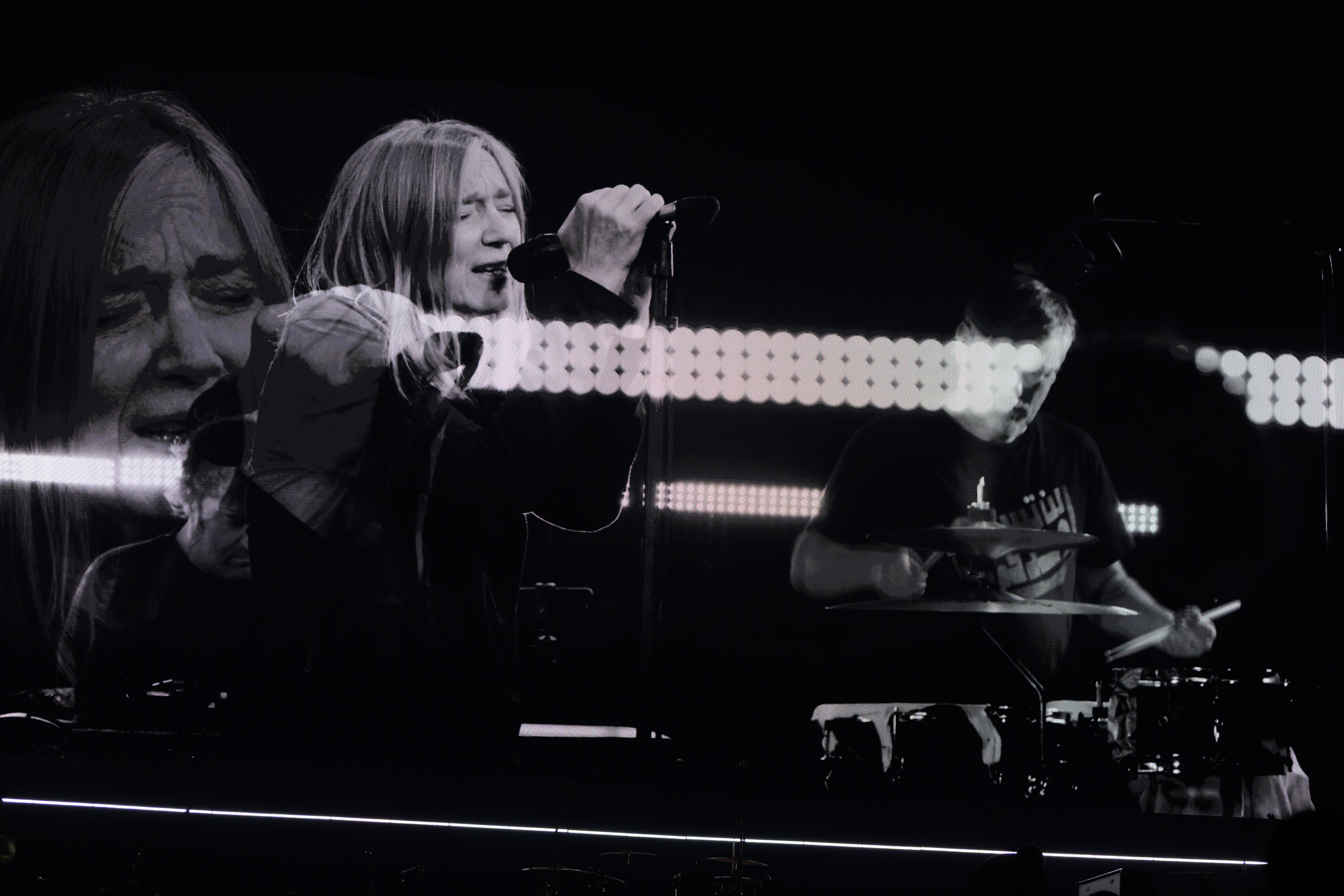Singer with long hair performing on stage, holding microphone, with drummer visible to left against dark background with stage lights.