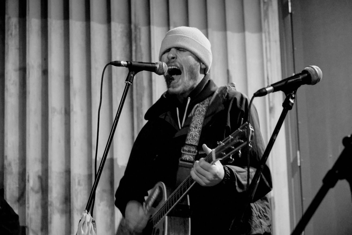 Man with beanie singing into microphone whilst playing guitar, mouth open, black and white image with curtained backdrop.