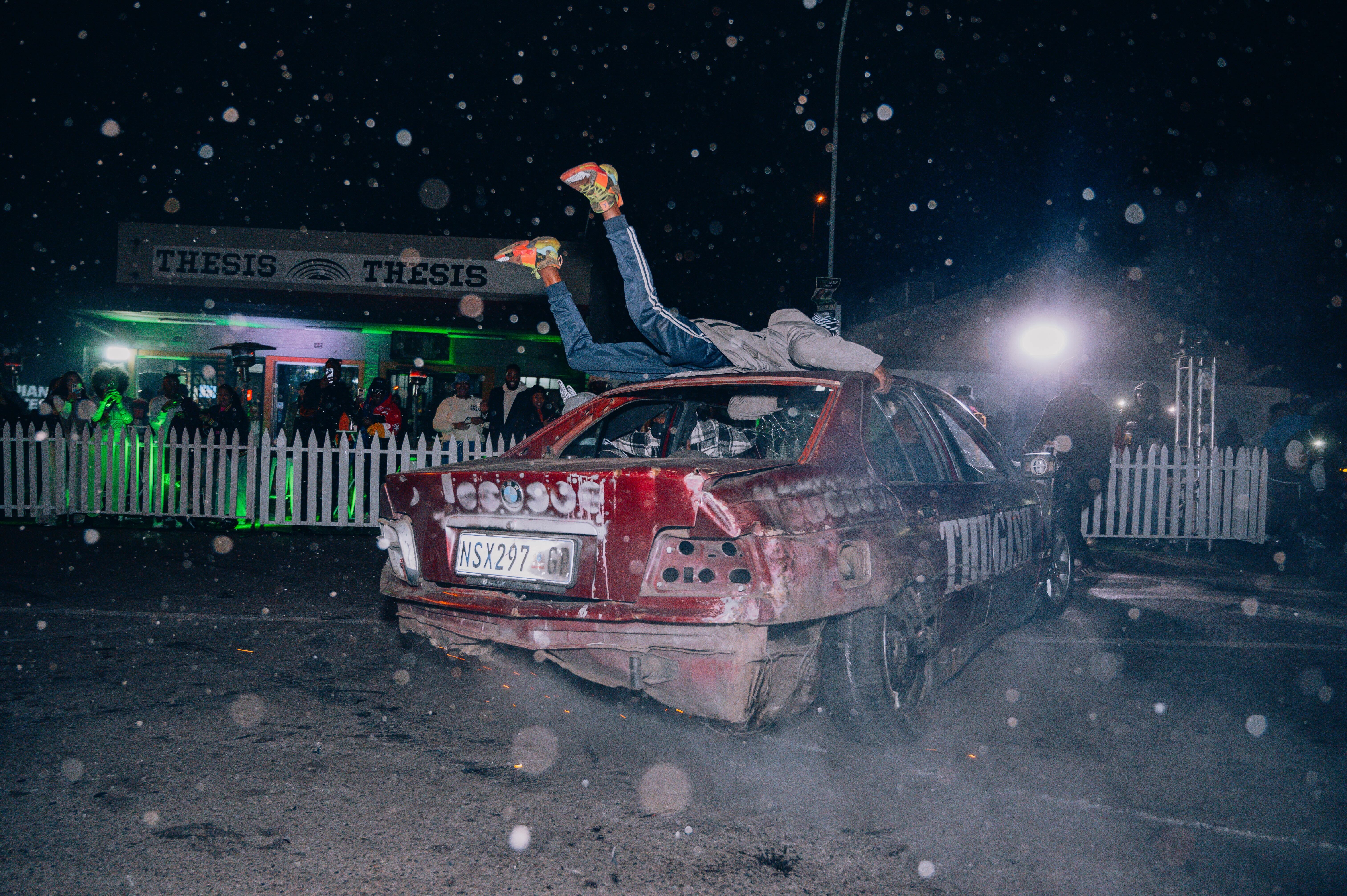 Crashed red car in a snowy scene at night, with green-lit 'Thesis' signs in the background.