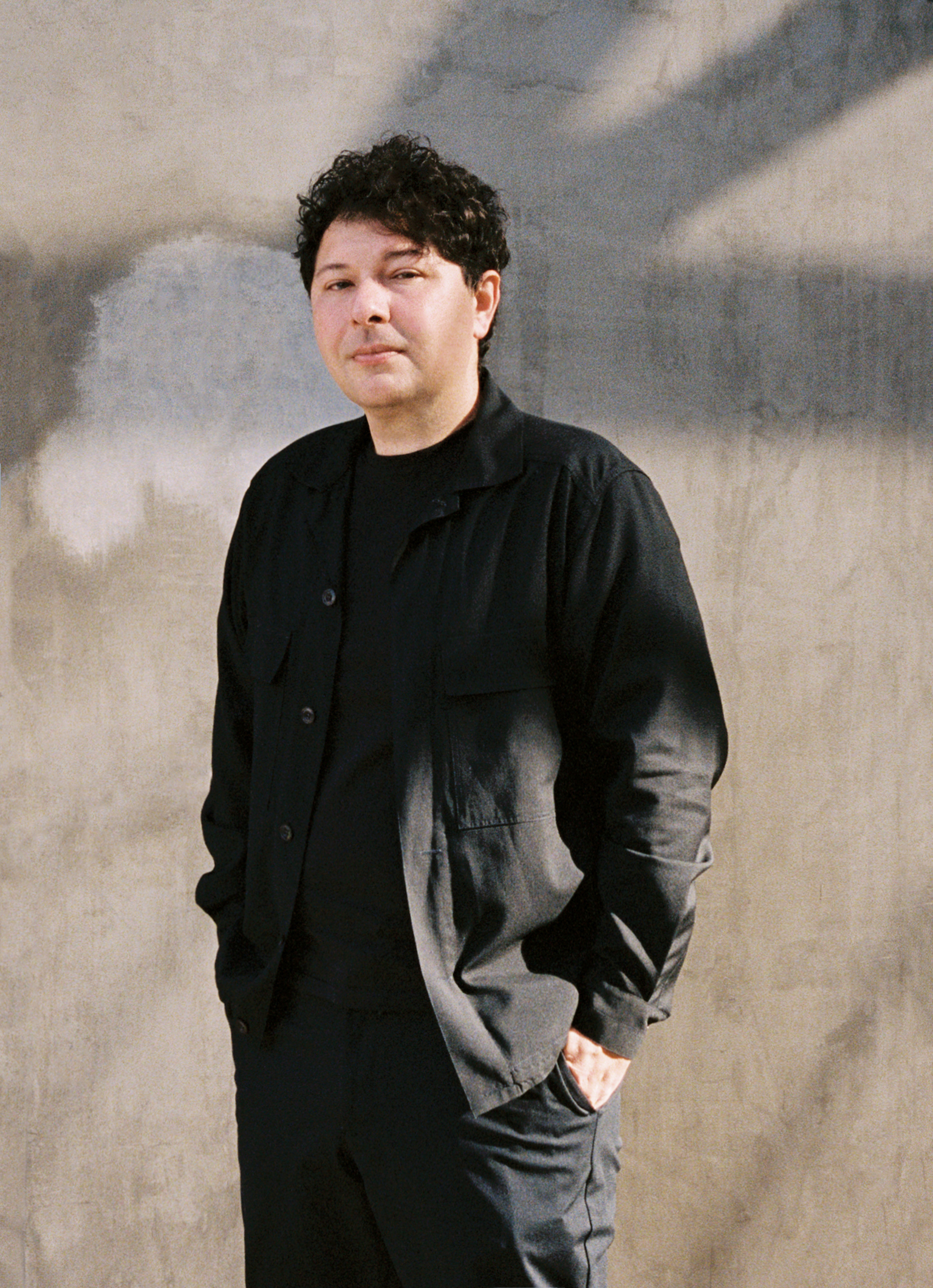 Person in black chef's jacket and trousers standing with hands in pockets against grey cloudy sky backdrop.