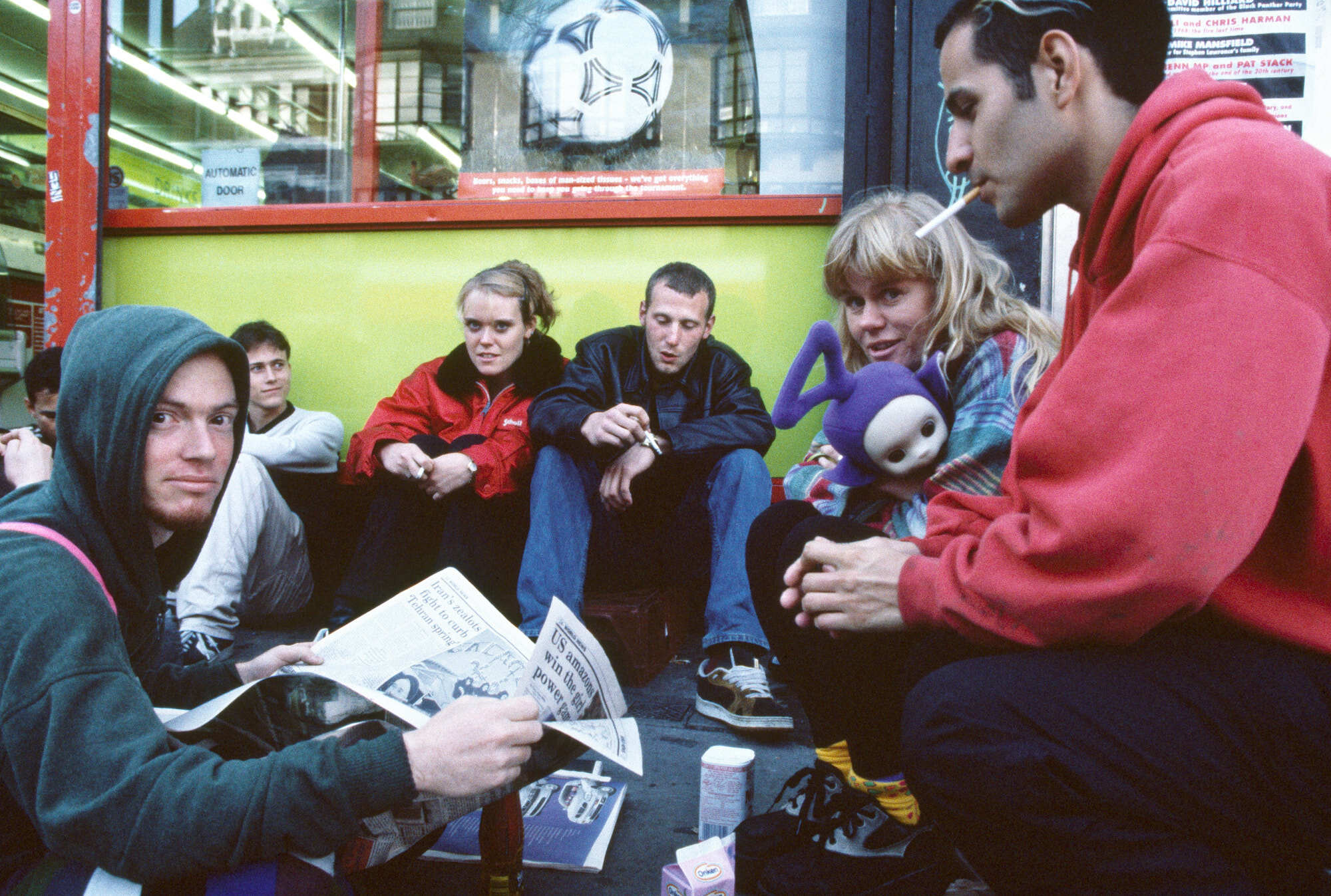 Six young people, three women and three men, sitting together in a brightly lit public space. They are engaged in casual conversation, some reading newspapers. The setting appears to be a train station or similar transport hub, with a large clock visible in the background.