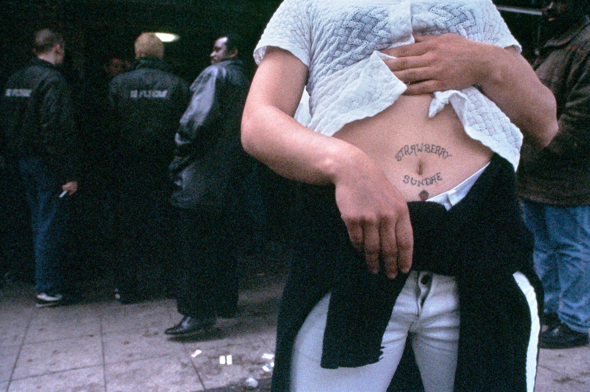 A person wearing a white T-shirt with a graphic design on their arm, standing in front of people in dark clothing.