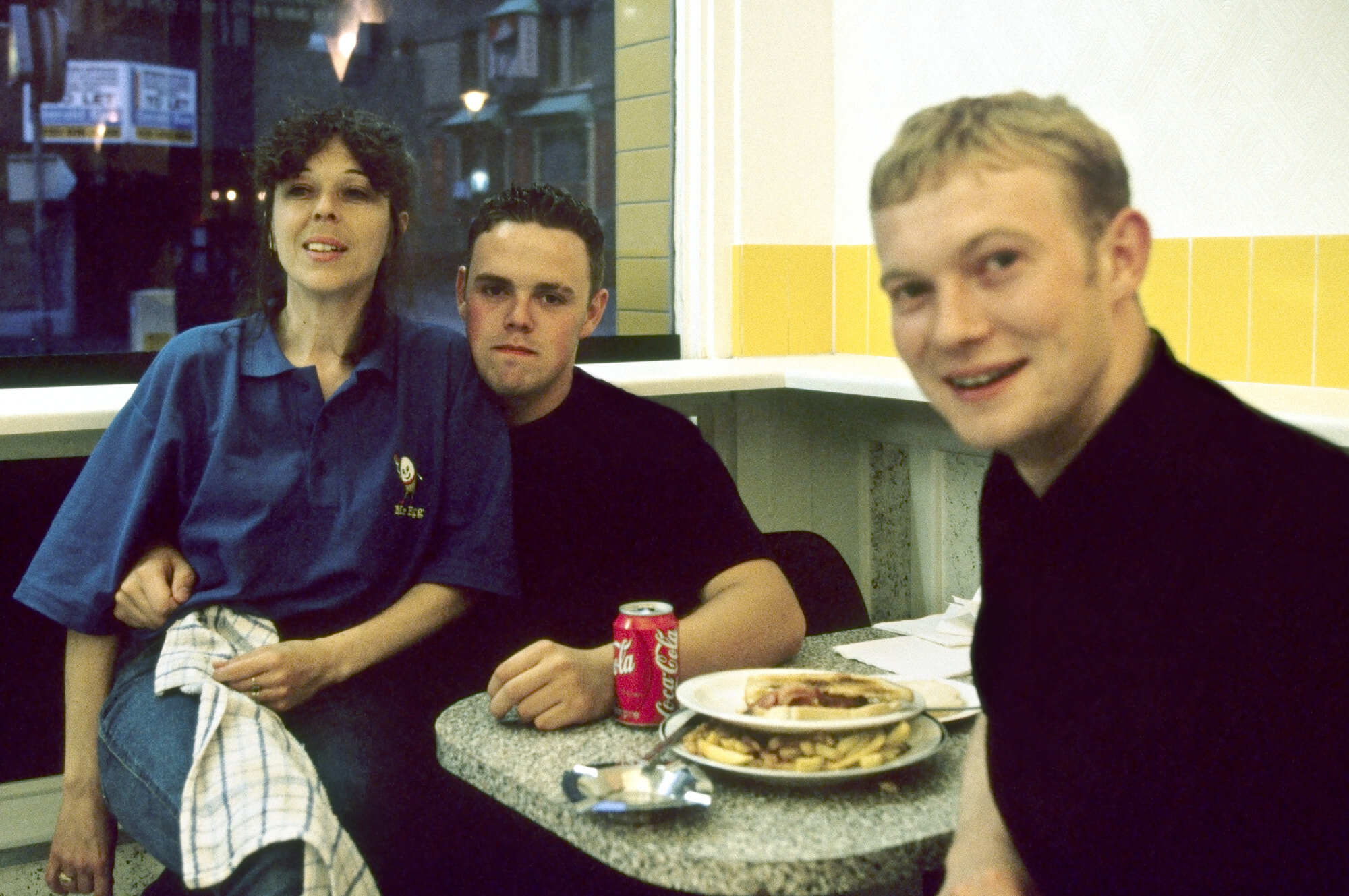 Three people sitting at a table in a diner, enjoying a meal and drinks. A woman in a blue shirt, a man in a black top, and another man in a black top, all smiling and engaged in conversation.