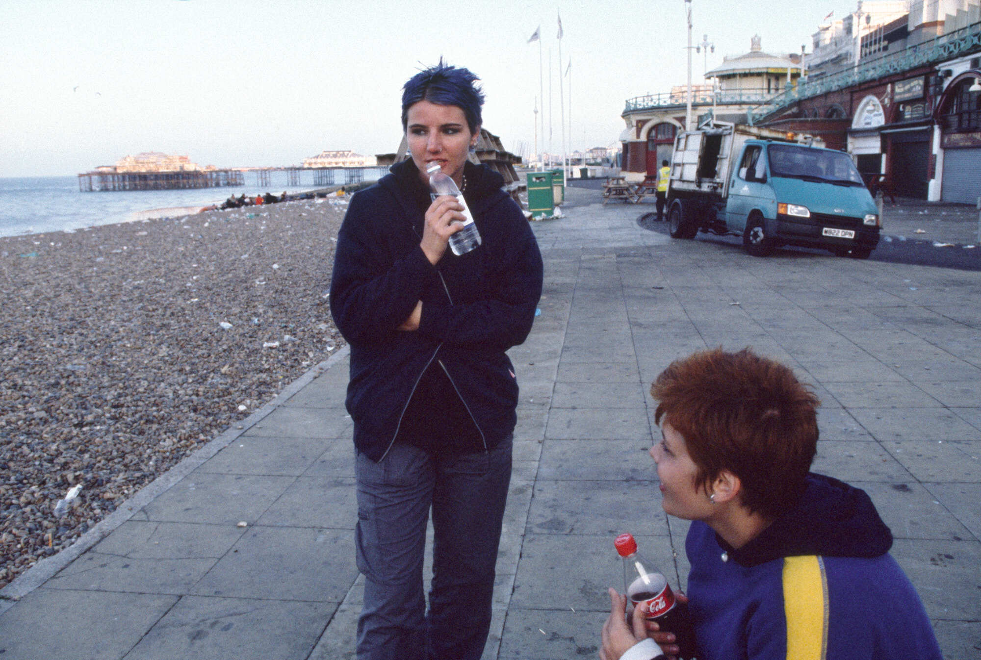 Couple standing on seaside promenade, woman holding a drink bottle, man holding a flower.