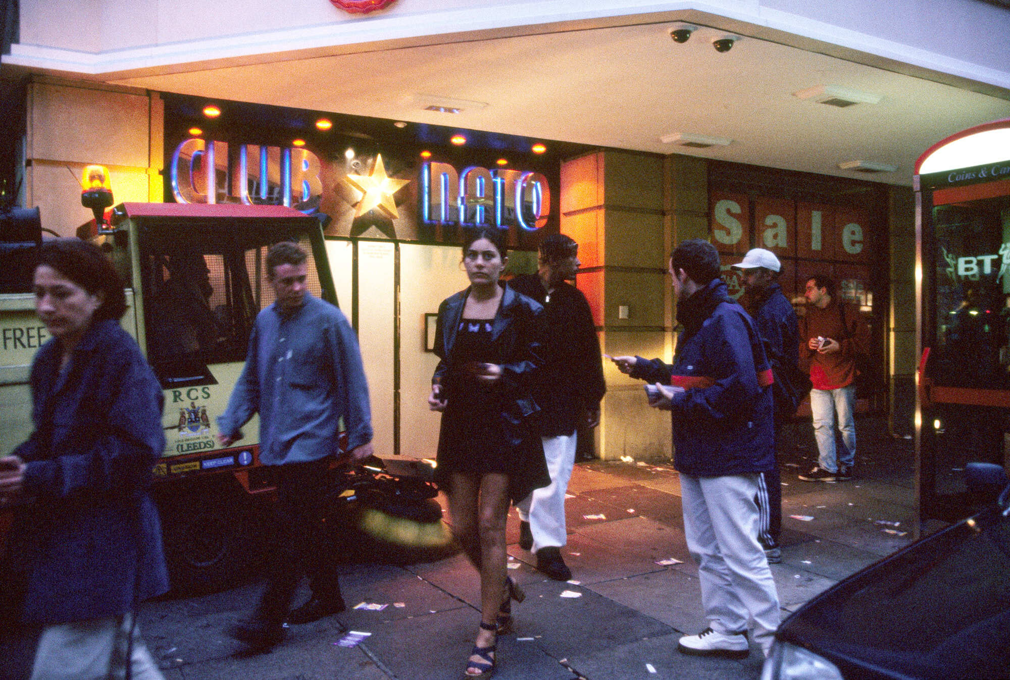 Crowded night scene with people walking on street, neon signs, and shop windows.