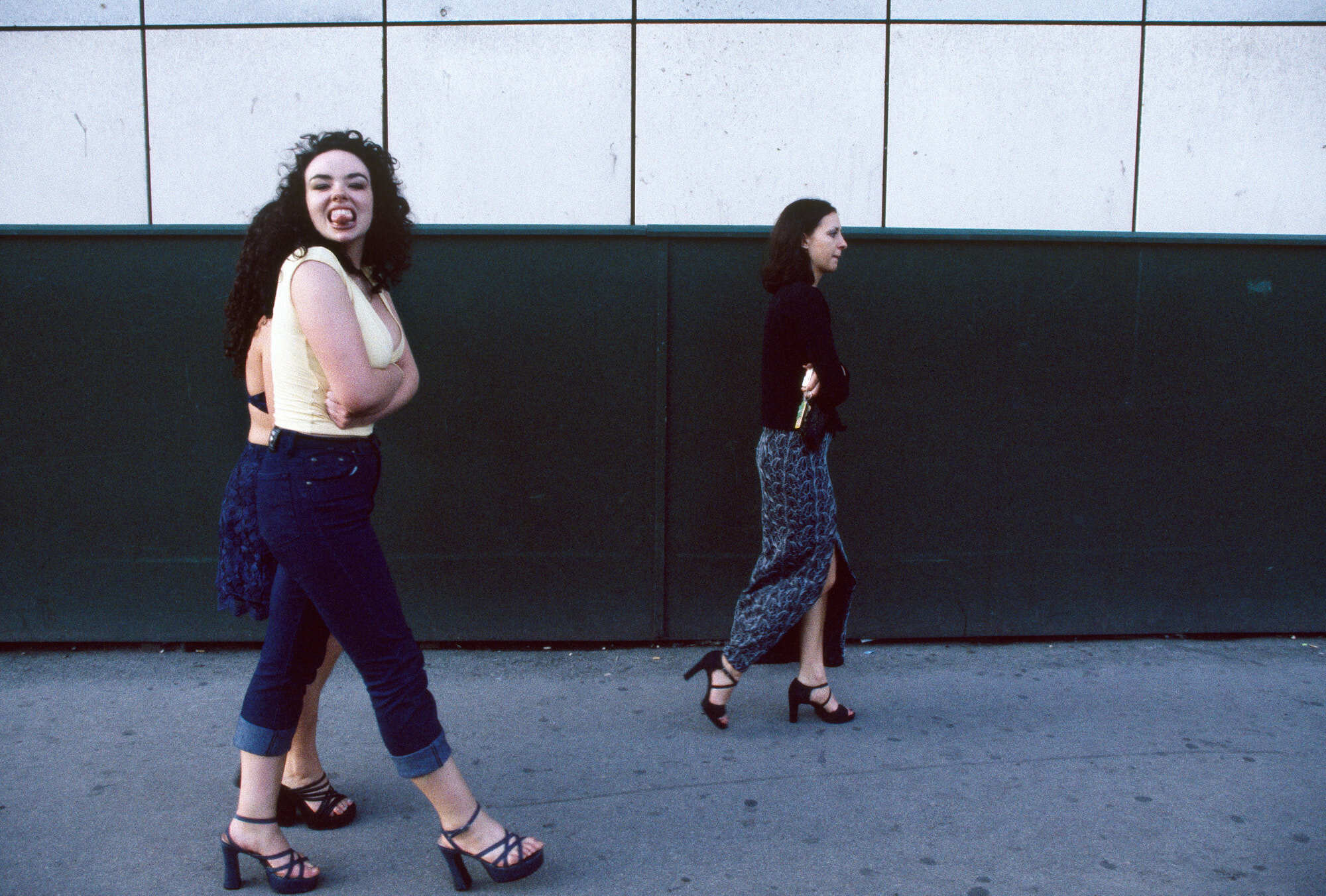 Two women standing against a dark wall, one in a pink top and the other in a burgundy dress, with their arms folded and legs crossed.