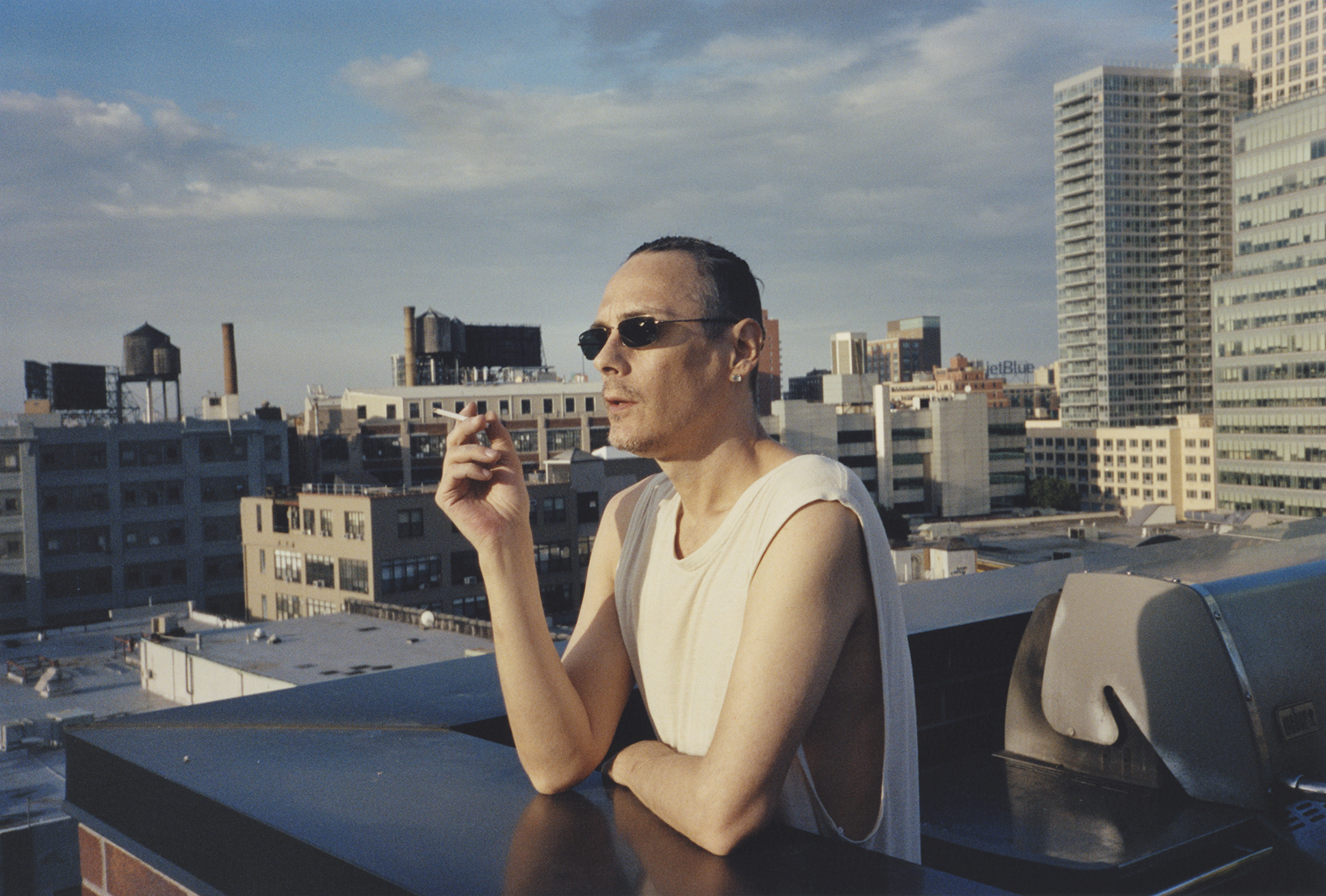 A person wearing sunglasses and a white vest stands on a balcony overlooking a city skyline.