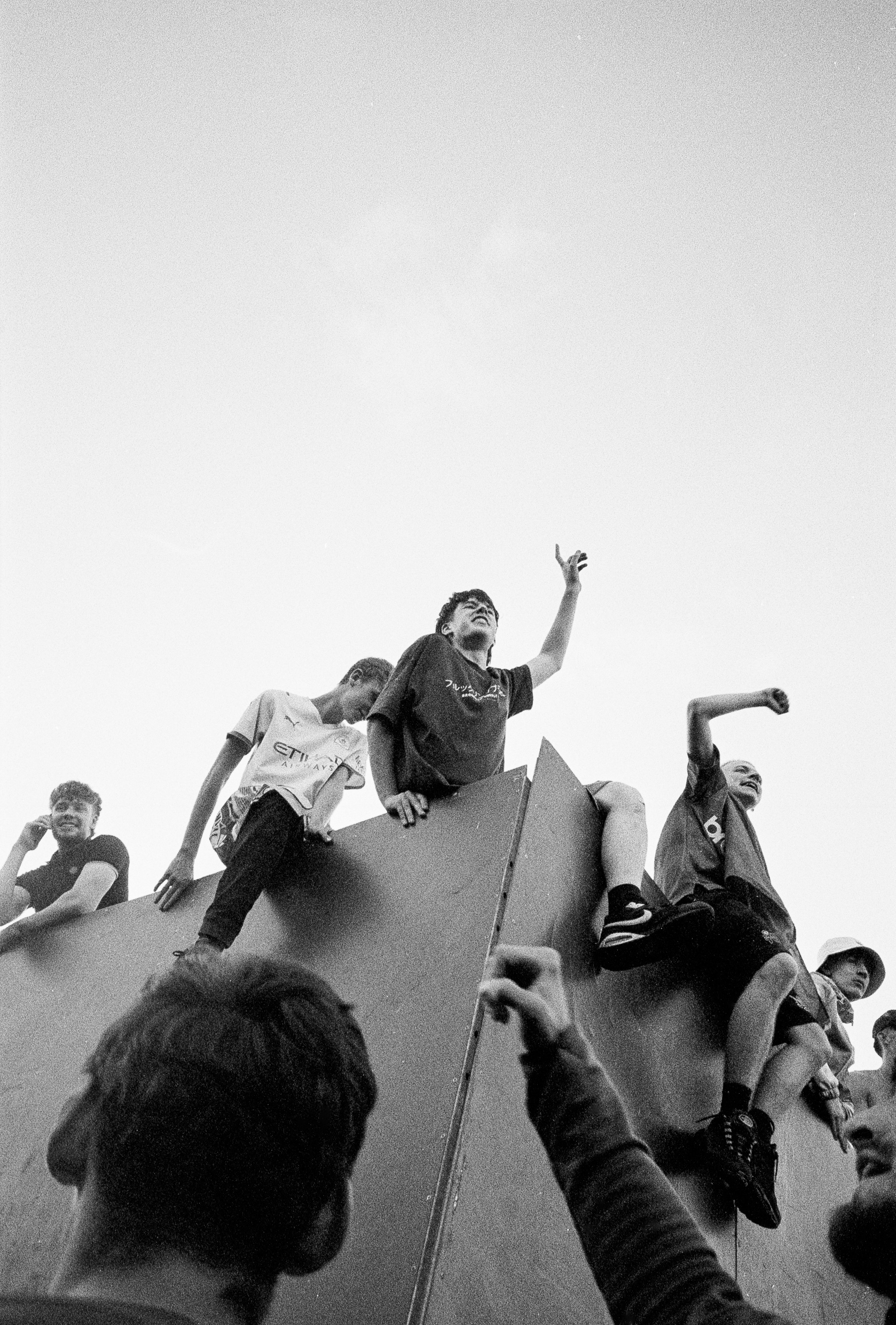 Black and white image showing people climbing and sitting on a large concrete structure, shot from below against overcast sky.
