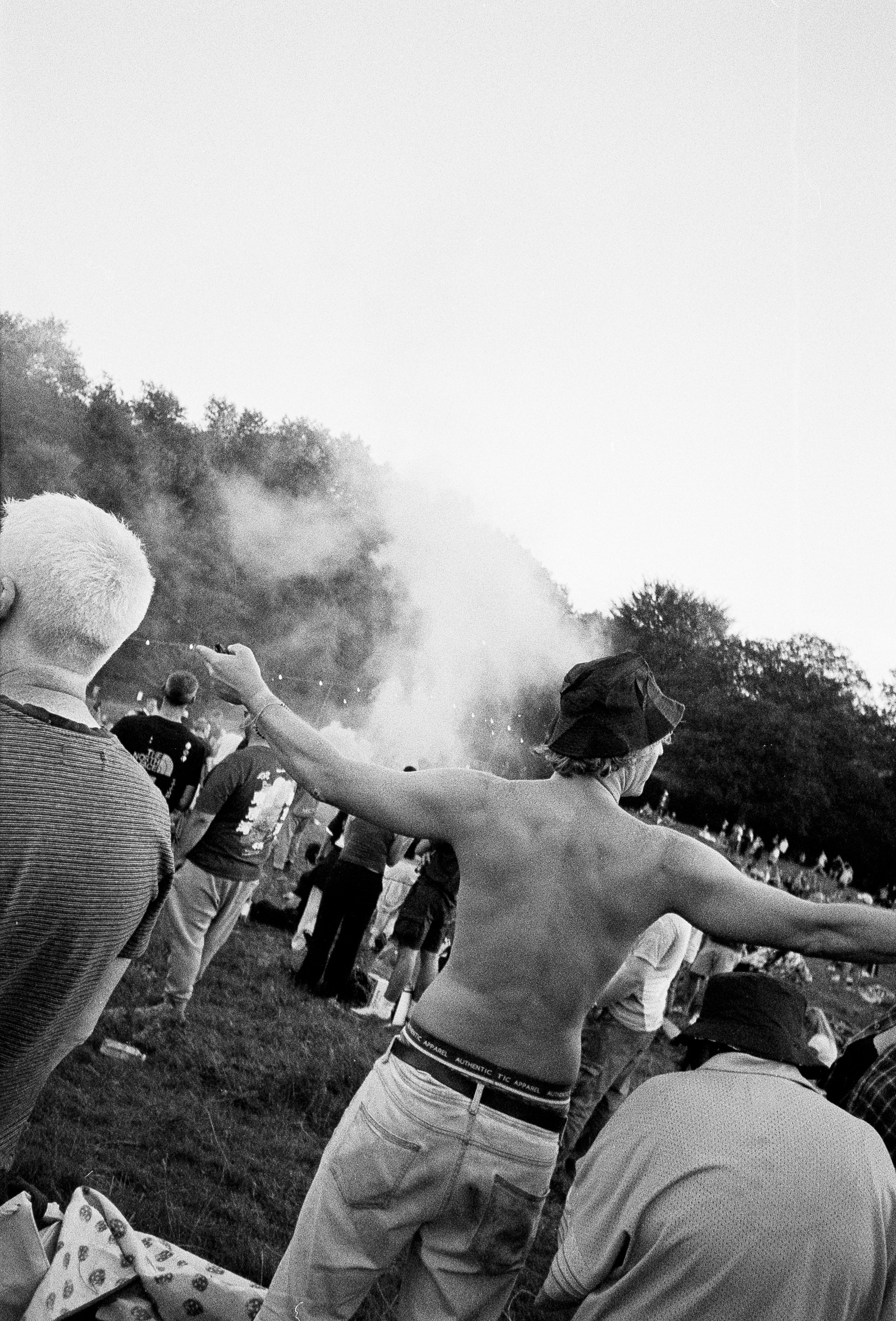 Shirtless man with arms raised spraying water or mist in crowded outdoor setting, black and white photograph with trees in background.